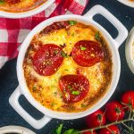 A baked dish in a white ramekin topped with three pepperoni slices, melted cheese, and sprinkled herbs, set on a dark surface near cherry tomatoes and a red-checkered cloth.