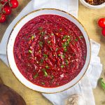 A bowl of vibrant red sauce garnished with chopped herbs sits on a white cloth, surrounded by tomatoes, garlic, and spices on a rustic yellow surface.