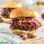 Two sloppy joe sandwiches on buns, topped with colorful coleslaw and pickle slices, sit on a white surface with scattered herbs and a few pieces of filling fallen out. A bowl of pickles is blurred in the background.