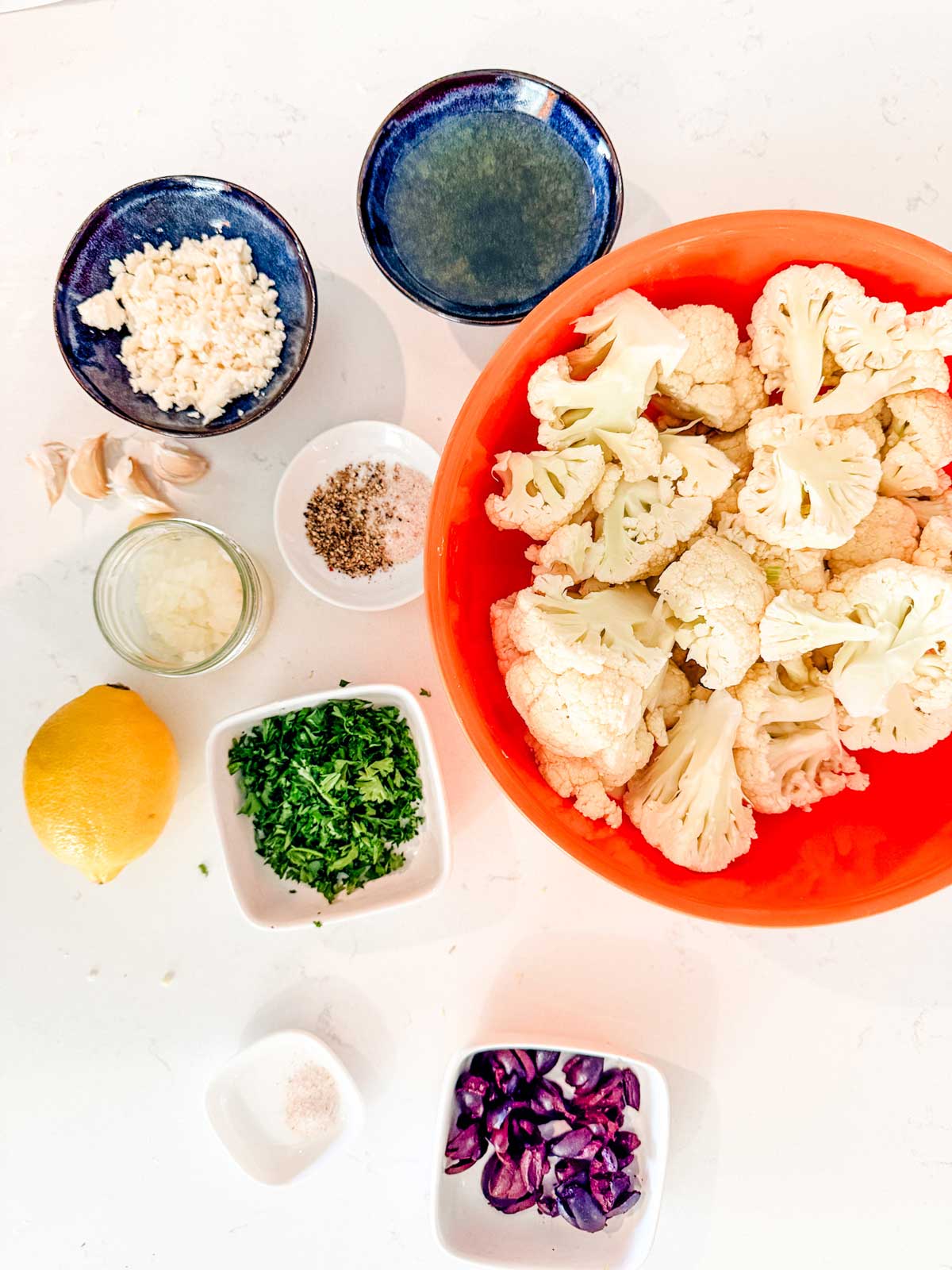 An overhead view of various ingredients on a white surface, including a large bowl of cauliflower florets, chopped herbs, lemon, minced garlic, chopped olives, spices, and small bowls with liquids and seasonings.