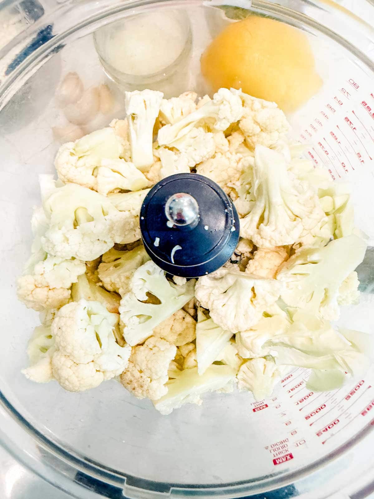 A food processor bowl filled with raw cauliflower florets, with part of a lemon and other ingredients visible in the background.
