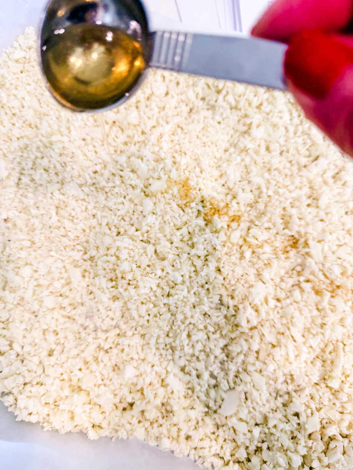 A hand holds a metal tablespoon of oil over a parchment lined sheet plan with finely chopped cauliflower.