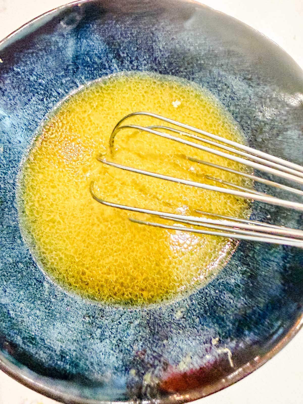 A metal whisk sits in a blue bowl containing a yellow, lemon dressing.
