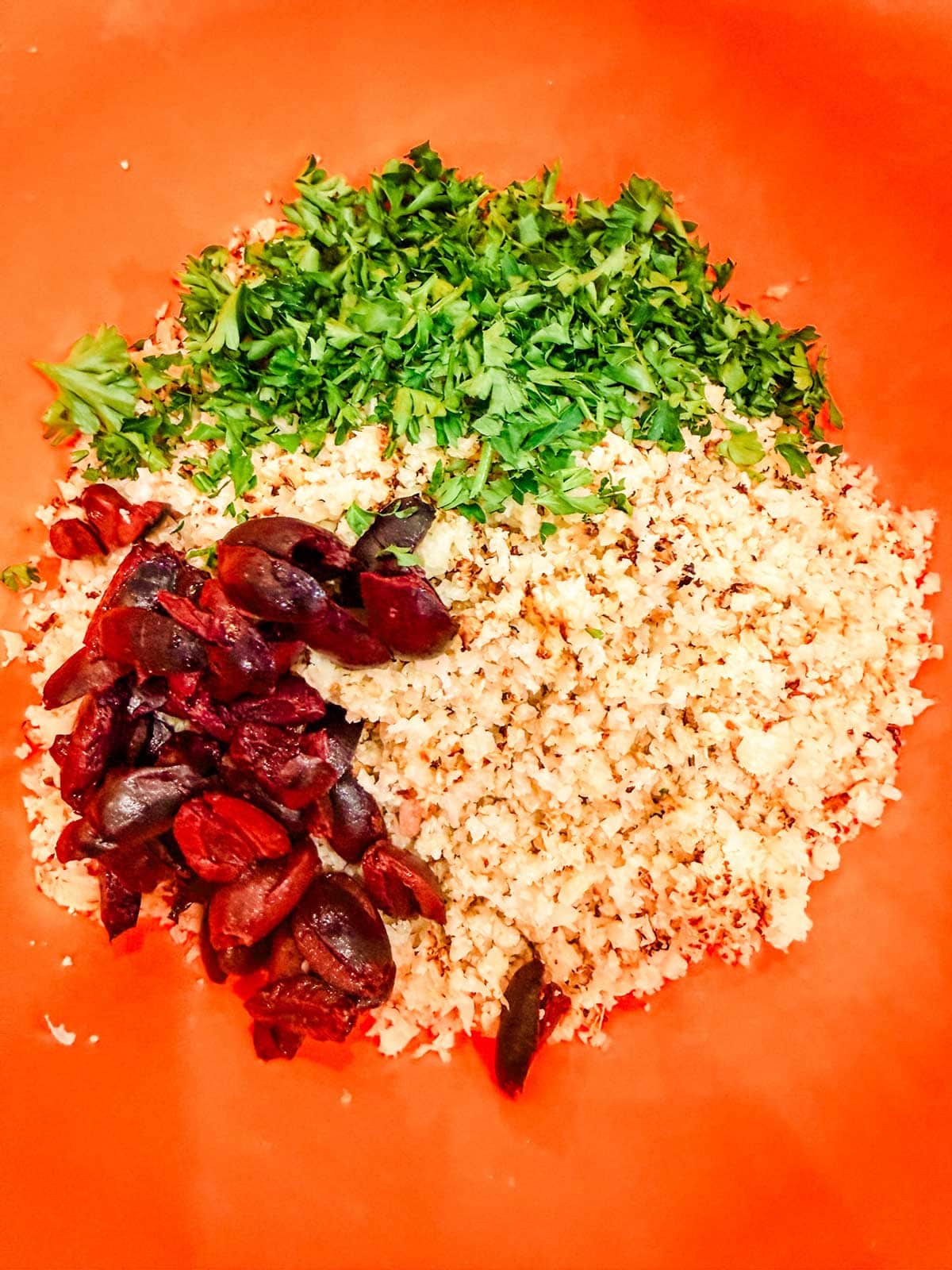 Chopped parsley, cauliflower rice, and sliced black olives arranged separately in an orange mixing bowl.