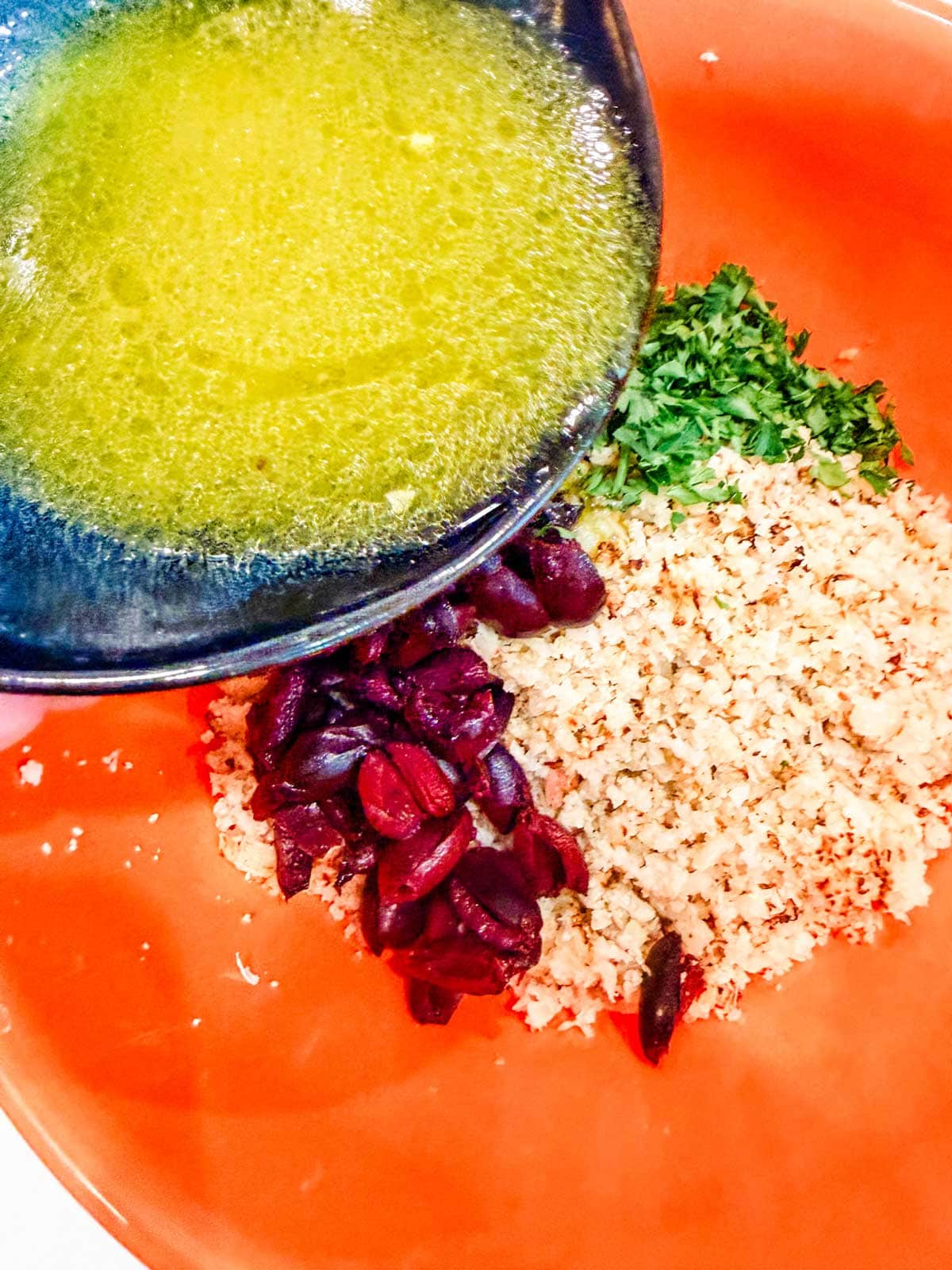Dressing being poured into a mixing bowl containing chopped parsley, cauliflower couscous, and sliced black olives on an orange surface.