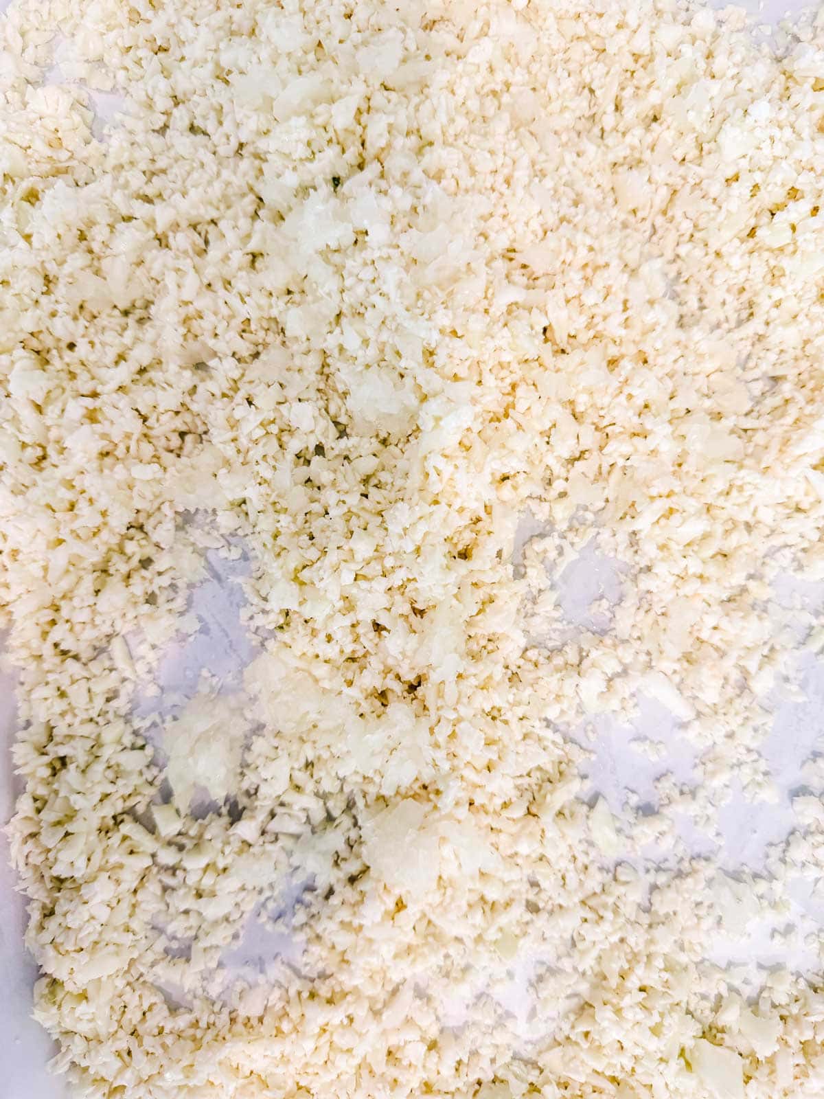 Close-up view of finely chopped or grated cauliflower spread out in a thin layer on a white surface, resembling couscous.