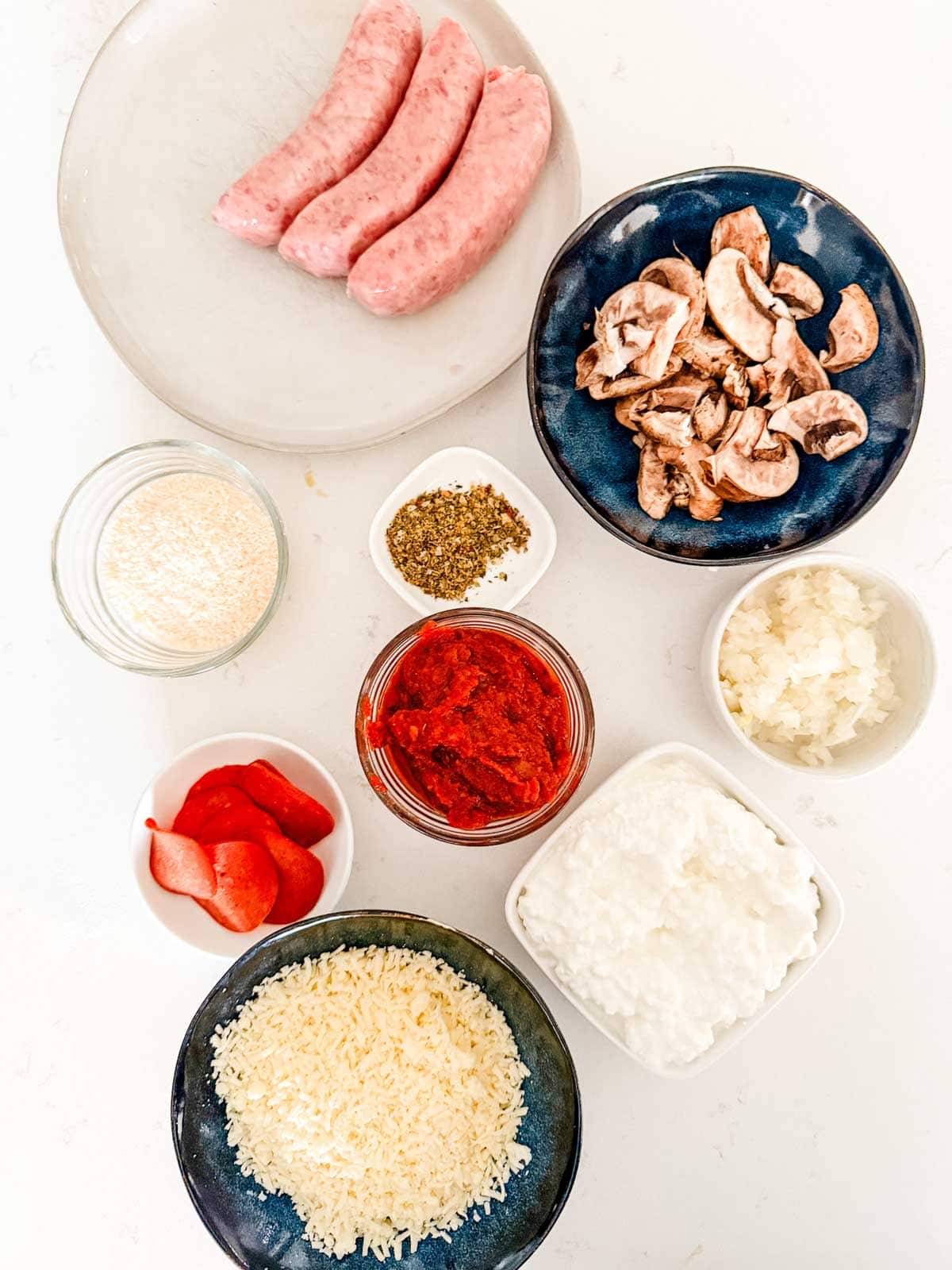 Flat lay of cooking ingredients on a white surface: raw sausages, sliced mushrooms, diced onions, tomato sauce, pepperoni, cottage cheese, shredded cheese, grated parmesan, and Italian seasoning in small bowls.
