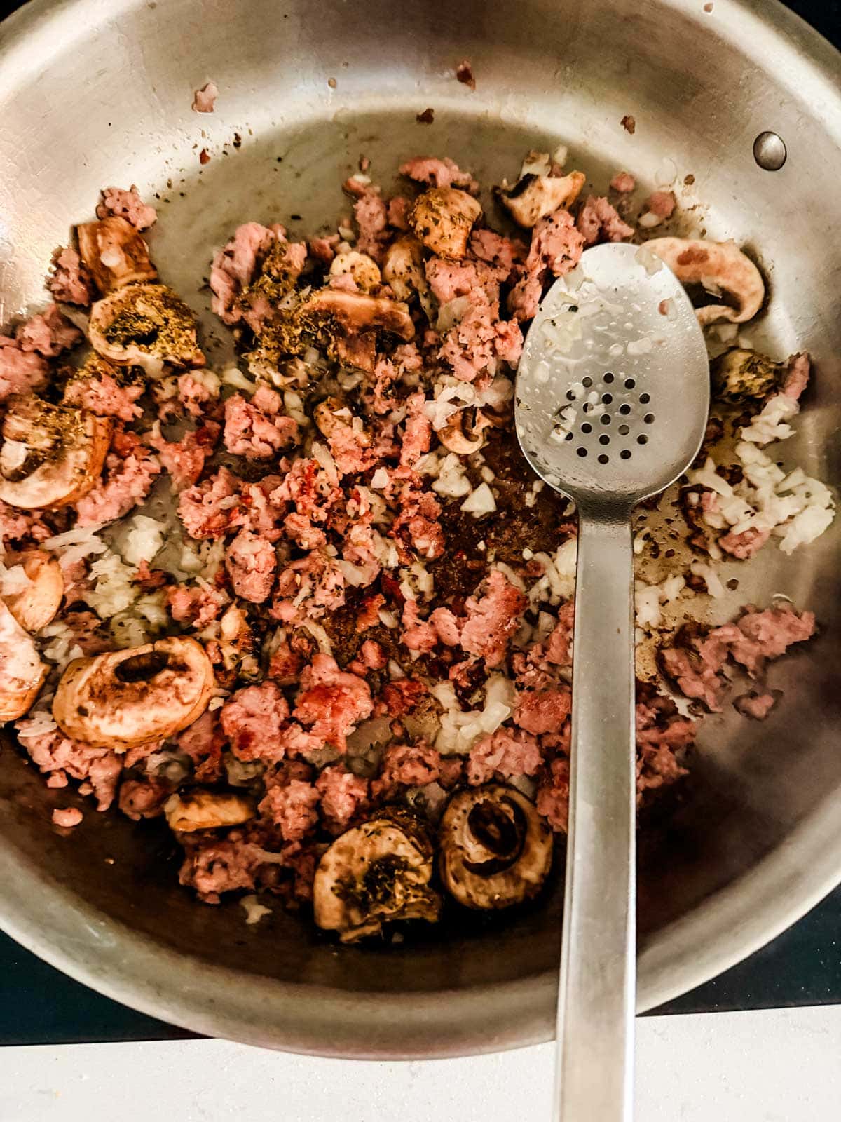 A metal pan with sausage, sliced mushrooms, chopped onions, and spices being sautéed. A slotted spoon rests inside the pan.
