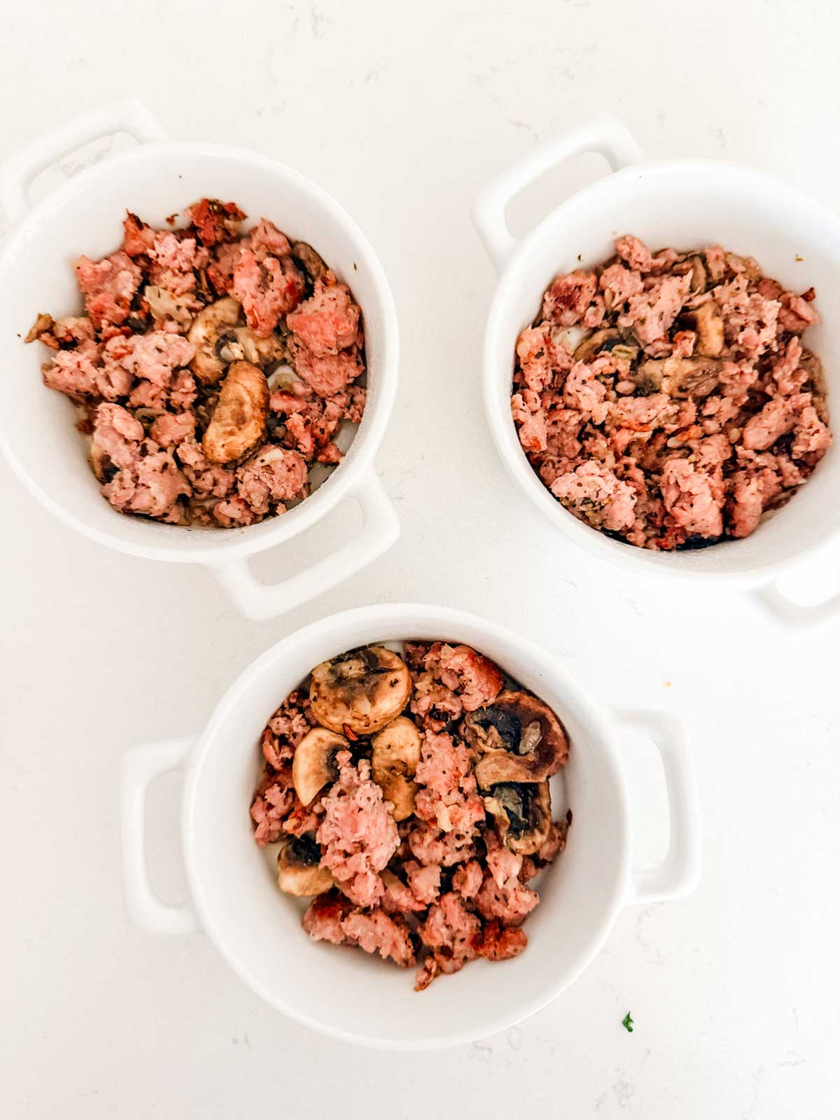 Three white ramekins filled with a mixture of cooked ground meat and mushrooms are arranged on a white surface.