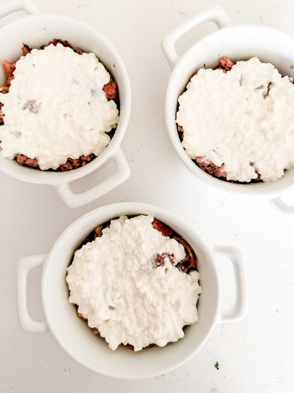 Three white ramekins filled with a layer of ground meat topped with a thick layer of cottage cheese, placed on a white surface.