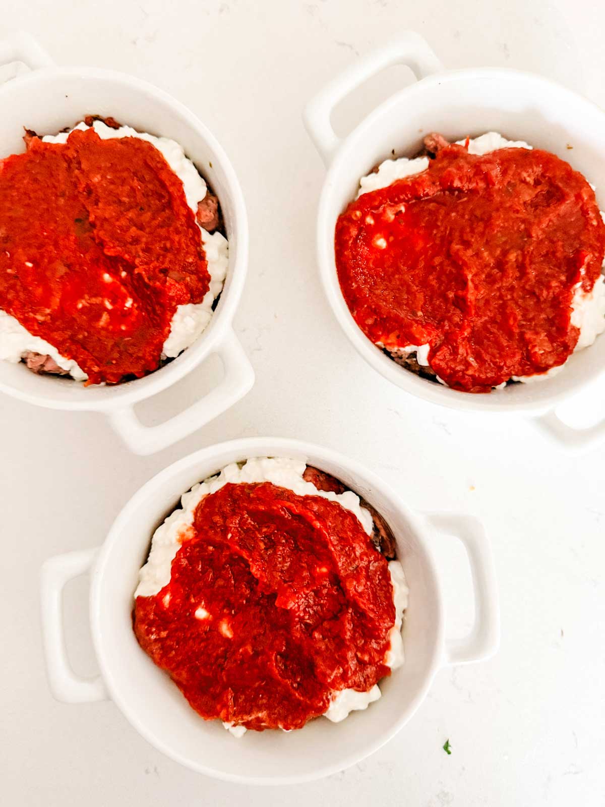 Three white ramekins filled with layers of ground meat, creamy white cheese, and topped with red tomato sauce, arranged on a white surface.