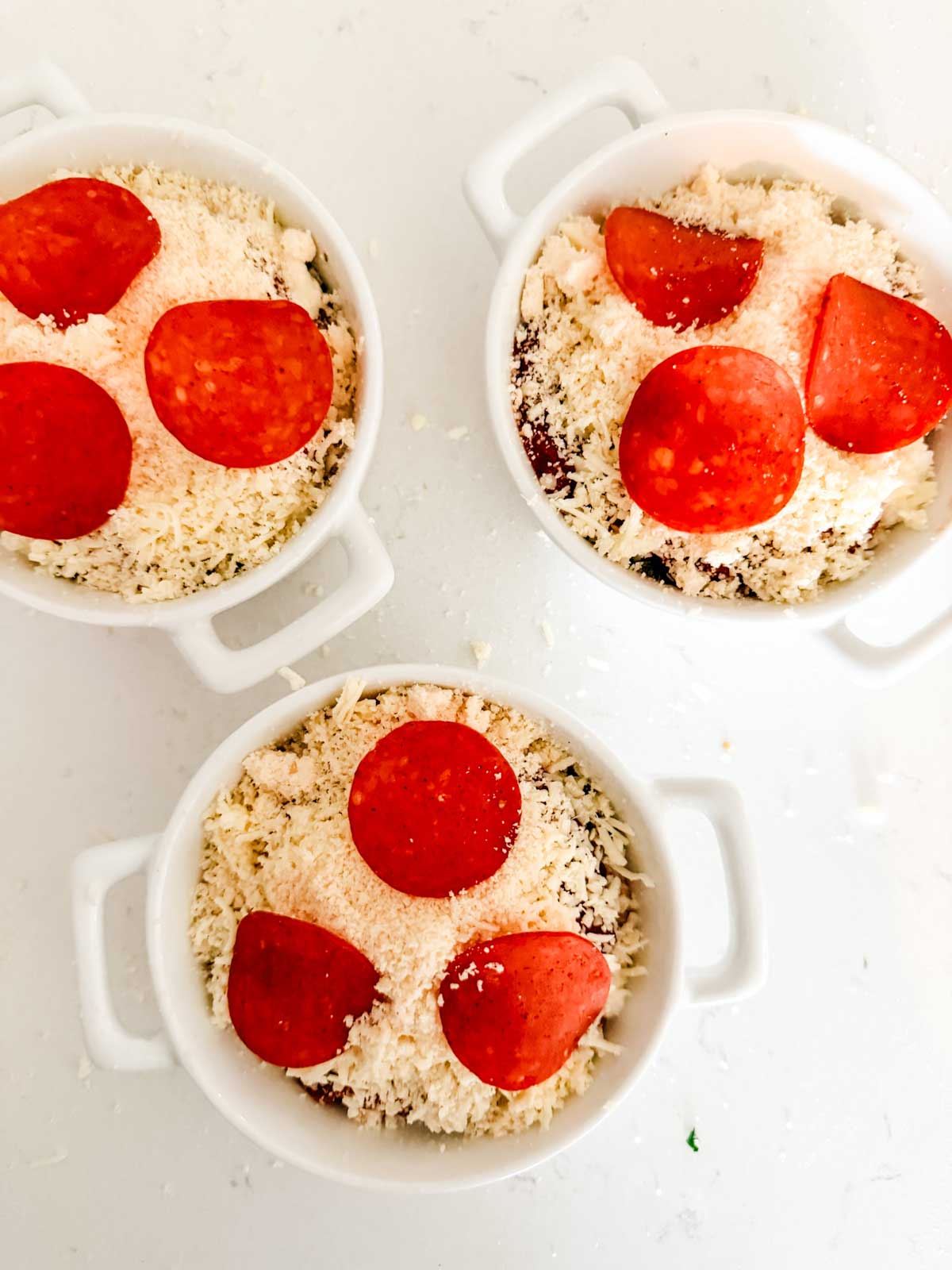 Three small white ramekins filled with shredded cheese and topped with three slices of pepperoni each, arranged on a white surface.