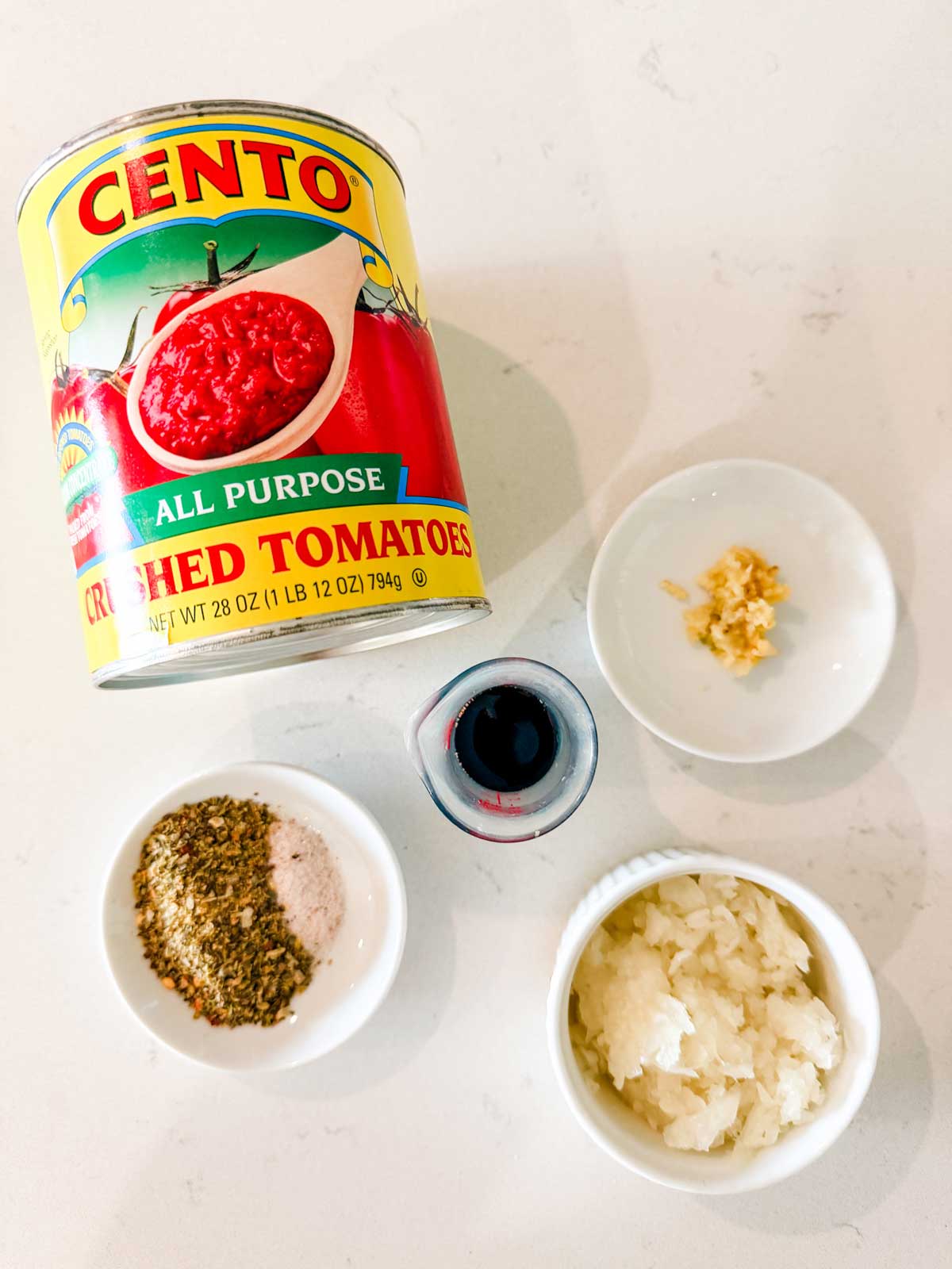 A can of Cento crushed tomatoes lies on its side next to small bowls containing minced garlic, finely chopped onion, dried herbs with salt, and a small cup of dark liquid on a white surface.