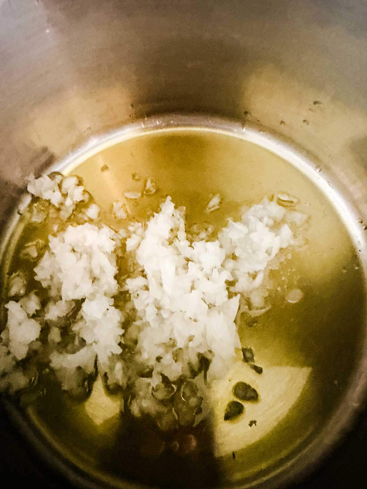 Finely chopped onions sautéing in oil at the bottom of a stainless steel pot.