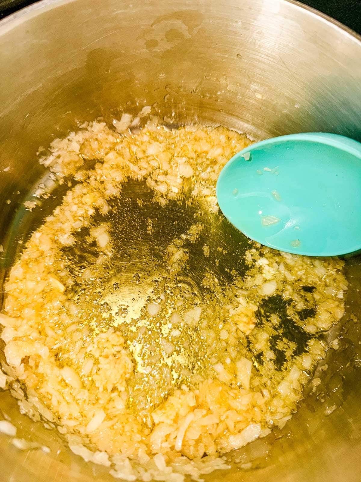 Chopped onions being sautéed in oil in a metal pot, with a blue spatula stirring the mixture.