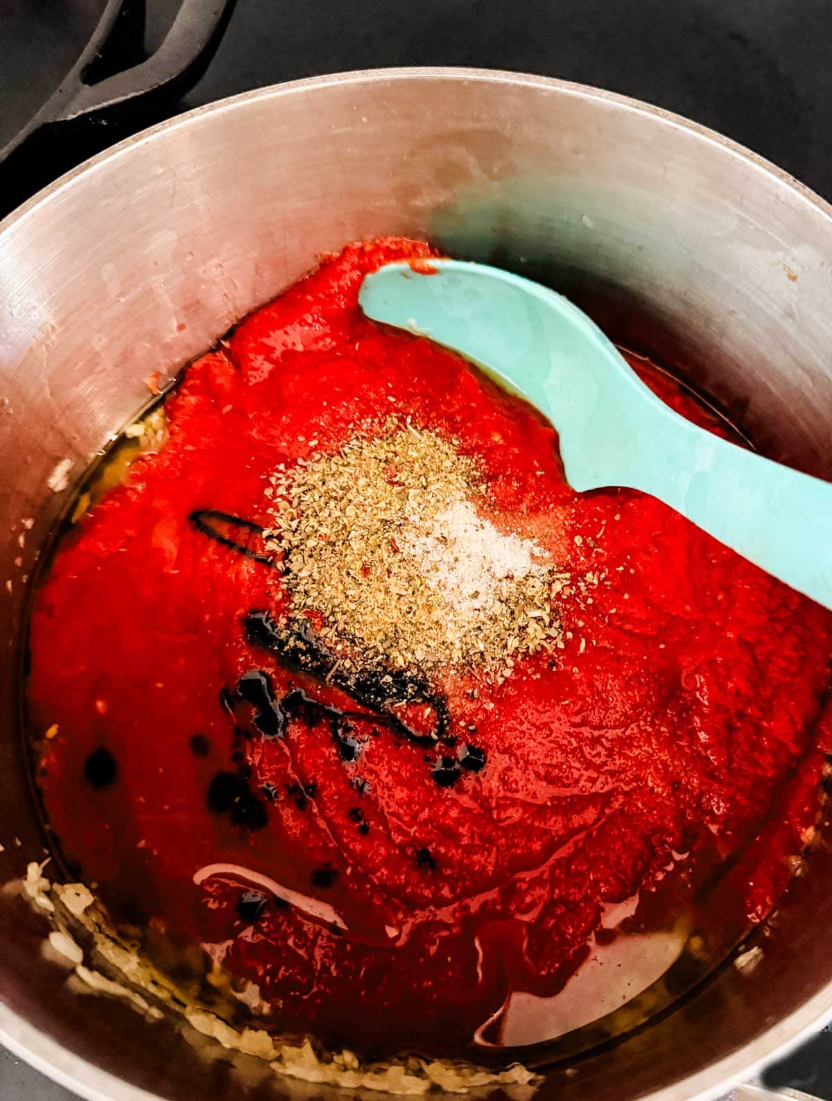 A metal pot filled with tomato sauce, minced garlic, herbs, spices, and a drizzle of olive oil. A blue silicone spoon rests in the pot, ready to stir the ingredients together.