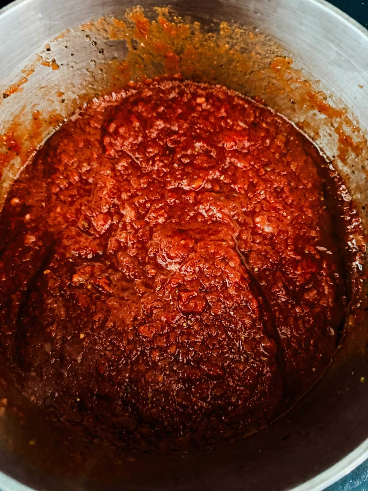 A close-up of a pot filled with thick, chunky red pizza sauce, simmering and reducing, with visible texture and oil on the surface.