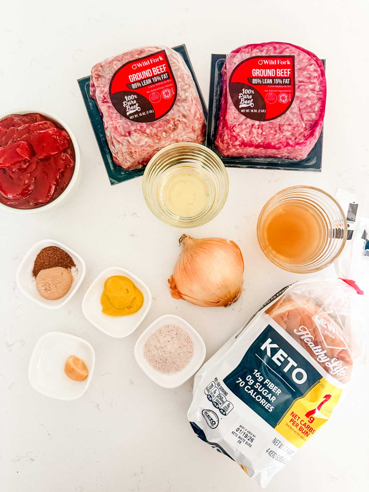 Ingredients for a recipe on a white surface: two packs of ground beef, a yellow onion, ketchup, mustard, spices, a clove of garlic, seasonings, broth, and package of keto buns.