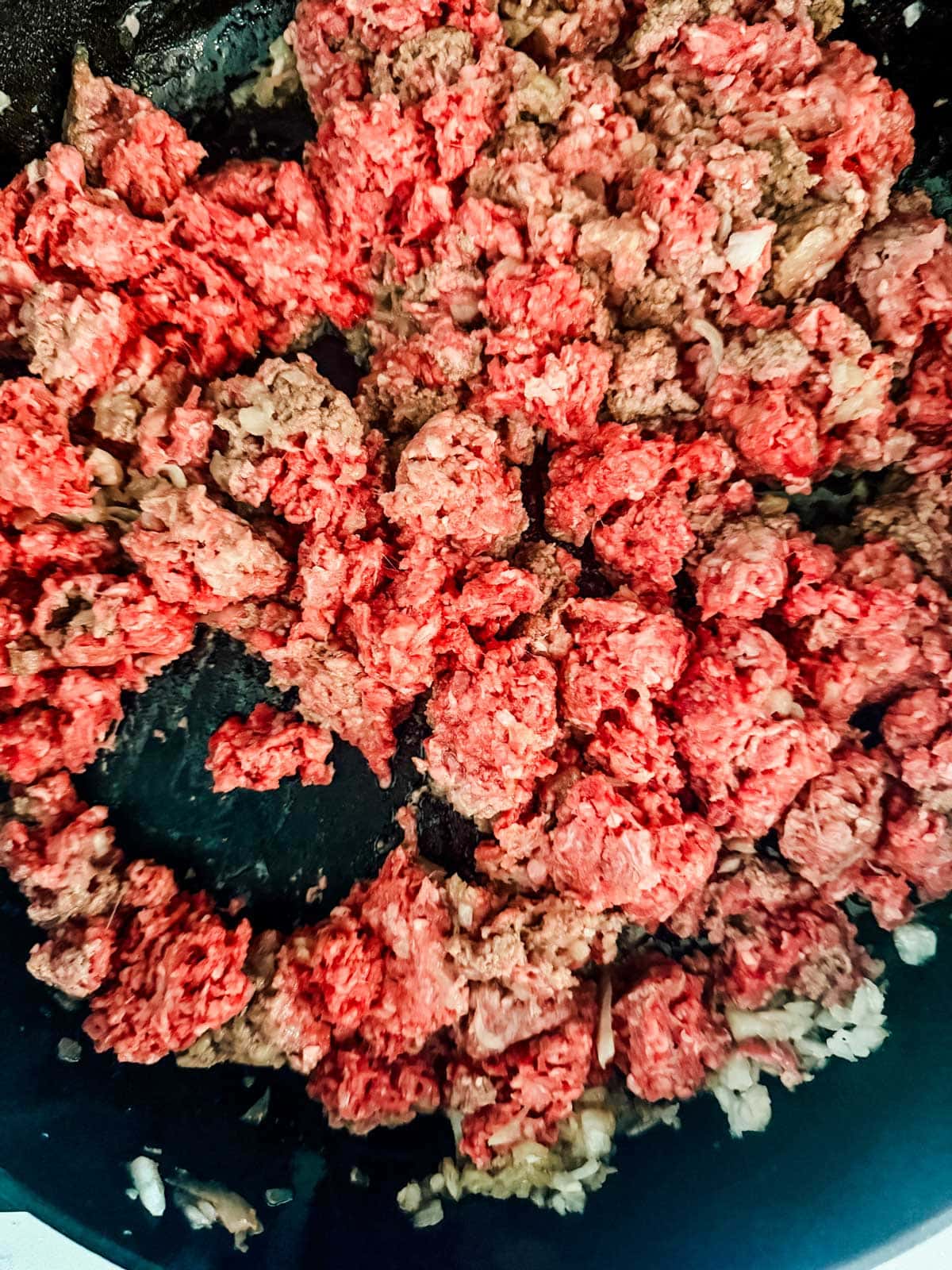 Ground beef being browned in a skillet with chopped onions. Some of the meat is still pink and uncooked, while other parts are starting to brown.