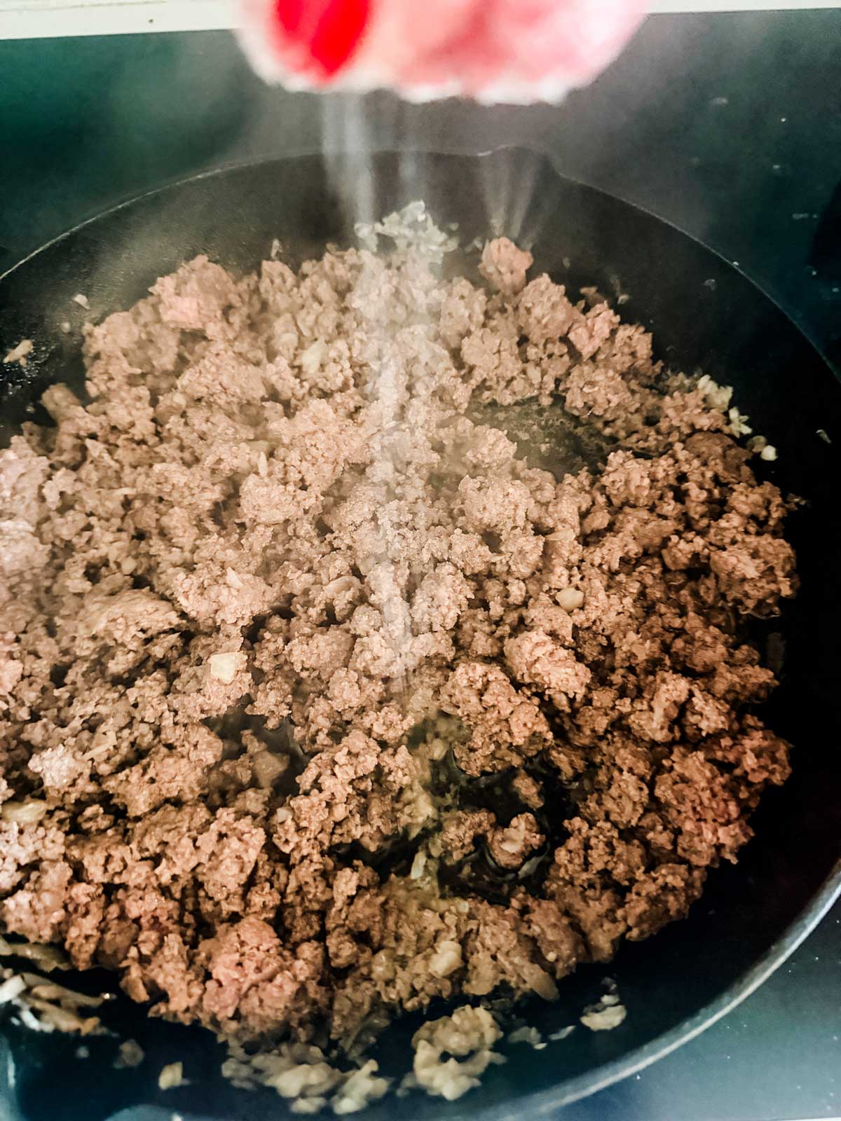 Ground beef and chopped onions are being cooked in a black skillet, with steam rising and salt being sprinkled from above.
