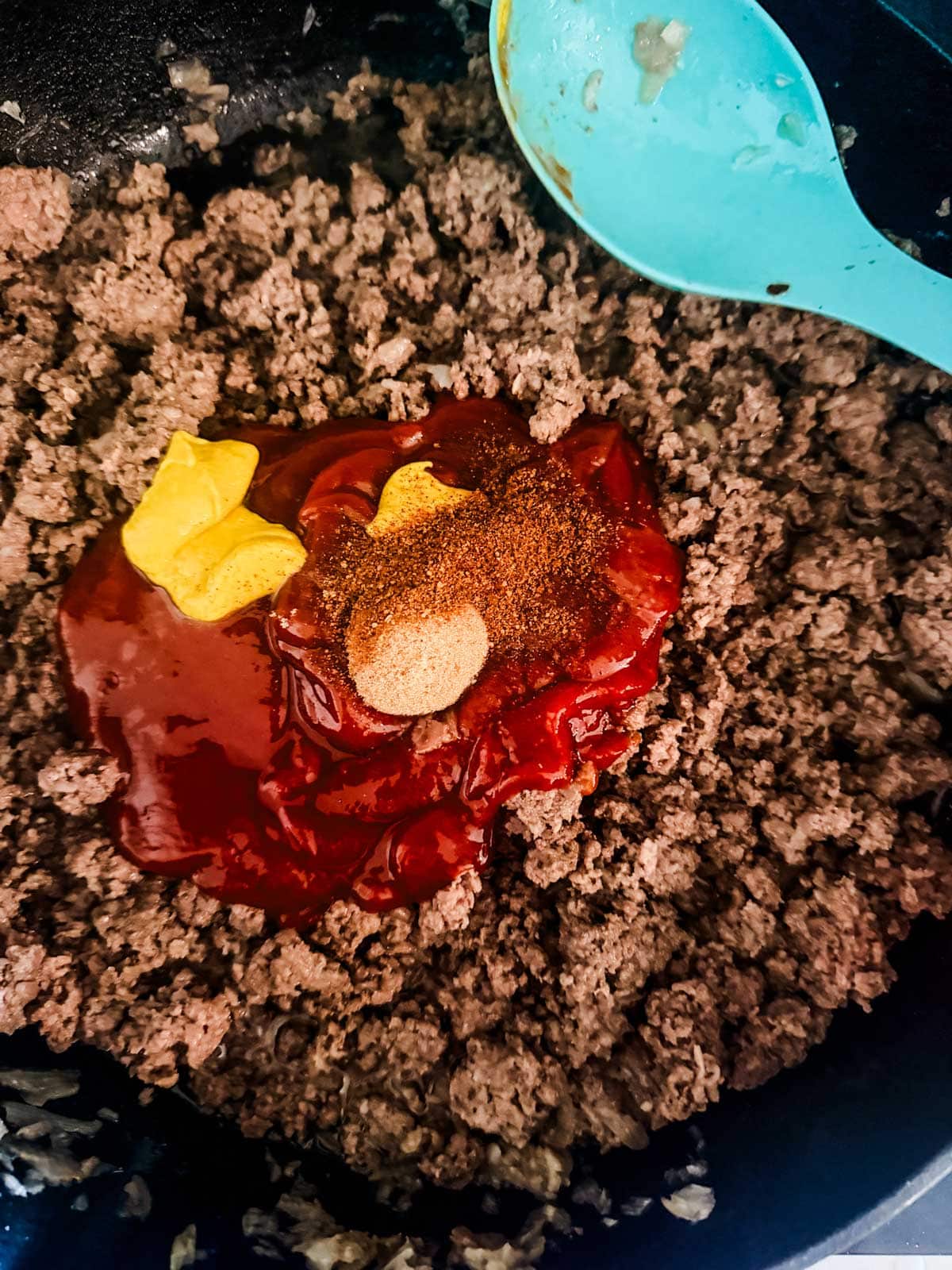 Ground beef cooking in a skillet topped with ketchup, mustard, spices, and a blue spoon resting on the side of the pan.