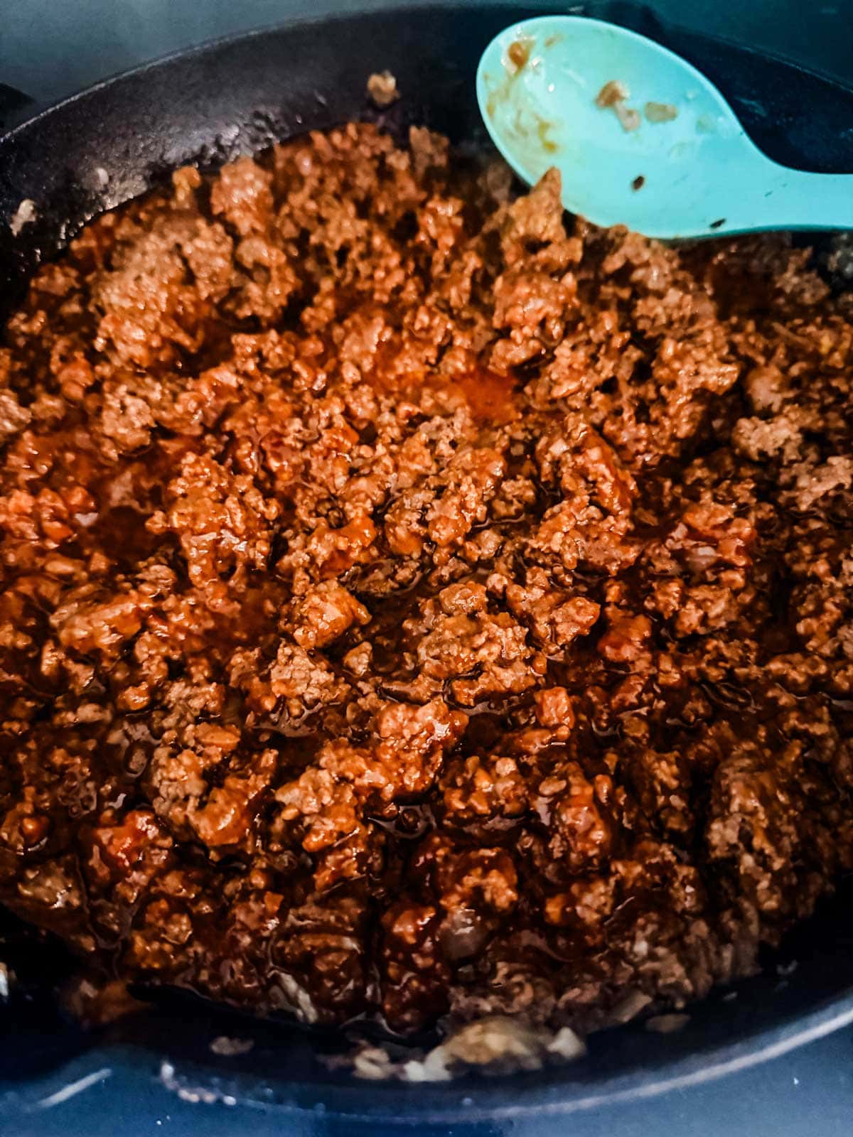 Ground beef cooked in a skillet with a rich, red sauce, and a blue spoon resting on the side.