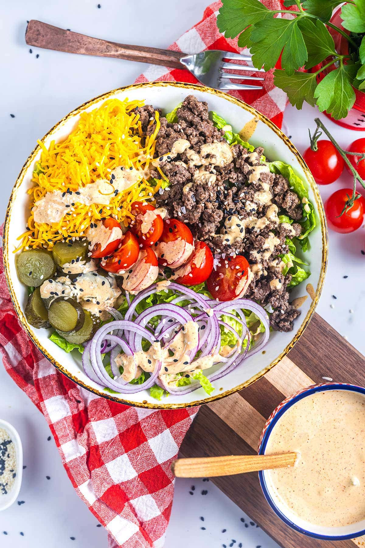 A keto Big Mac bowl filled with ground beef, shredded cheese, sliced pickles, cherry tomatoes, red onion, and lettuce, drizzled with sauce. Served with a red checkered napkin, fresh parsley, tomatoes, and dressing on the side.