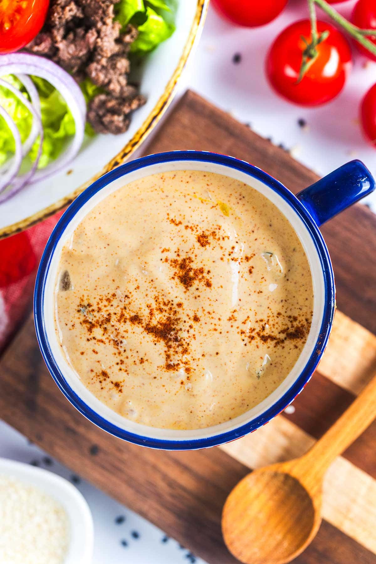A blue-rimmed mug filled with creamy, orange-colored sauce topped with a sprinkle of paprika sits on a wooden board next to a wooden spoon. Nearby are fresh tomatoes and a bowl of salad with onions and ground beef.