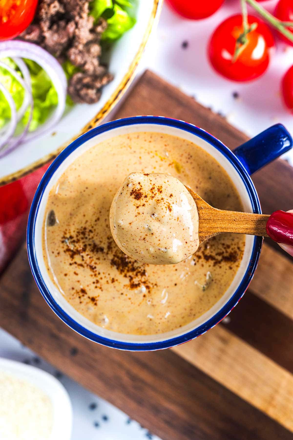 A wooden spoon lifts creamy, seasoned sauce from a blue-rimmed cup, with fresh vegetables and ground beef visible in the background.