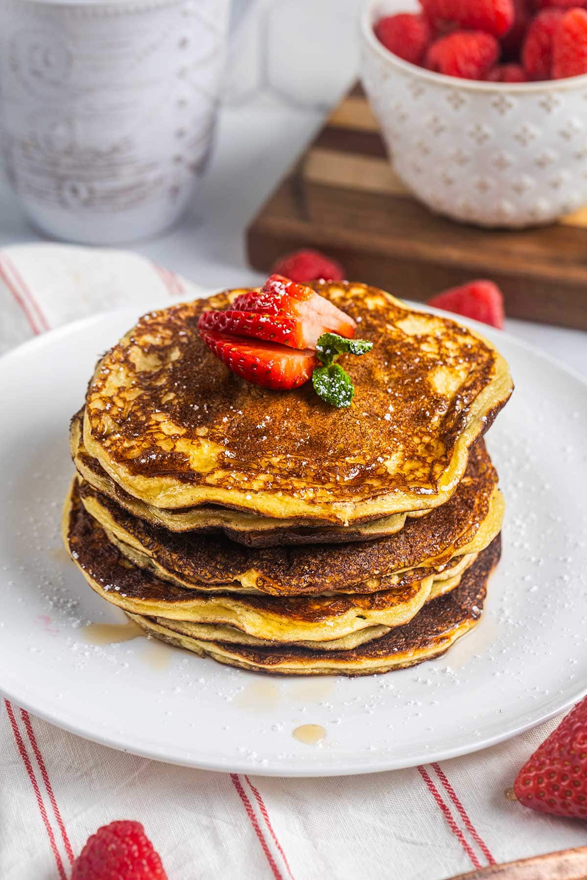 A stack of pancakes topped with sliced strawberries and a sprig of mint, served on a white plate with syrup. A bowl of strawberries, a wooden board, and a keto Big Mac bowl are in the background.