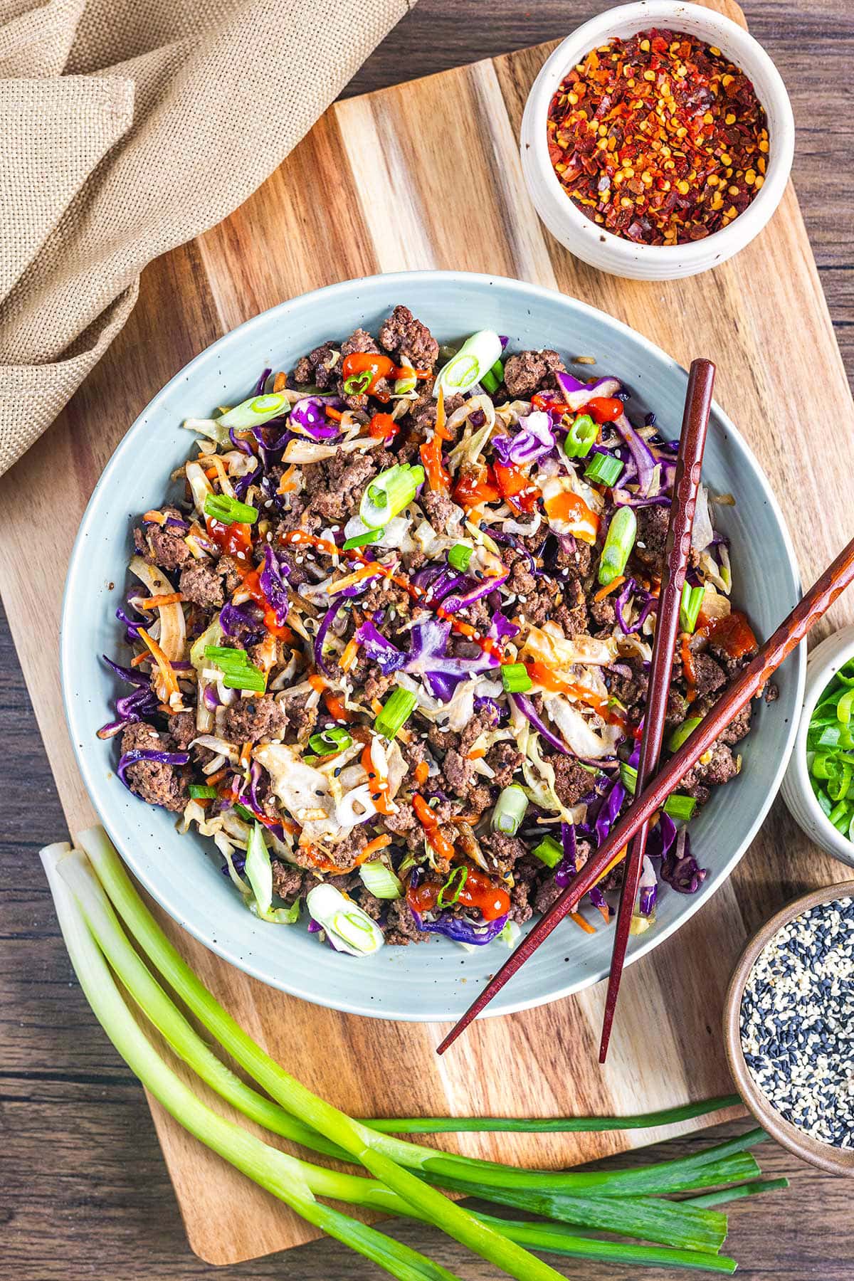 Overhead view of a keto egg roll in a bowl&mdash;stir-fried beef, shredded cabbage, green onions, and carrots topped with sesame seeds and chili sauce. Wooden chopsticks rest on the bowl with condiments and fresh green onions nearby.