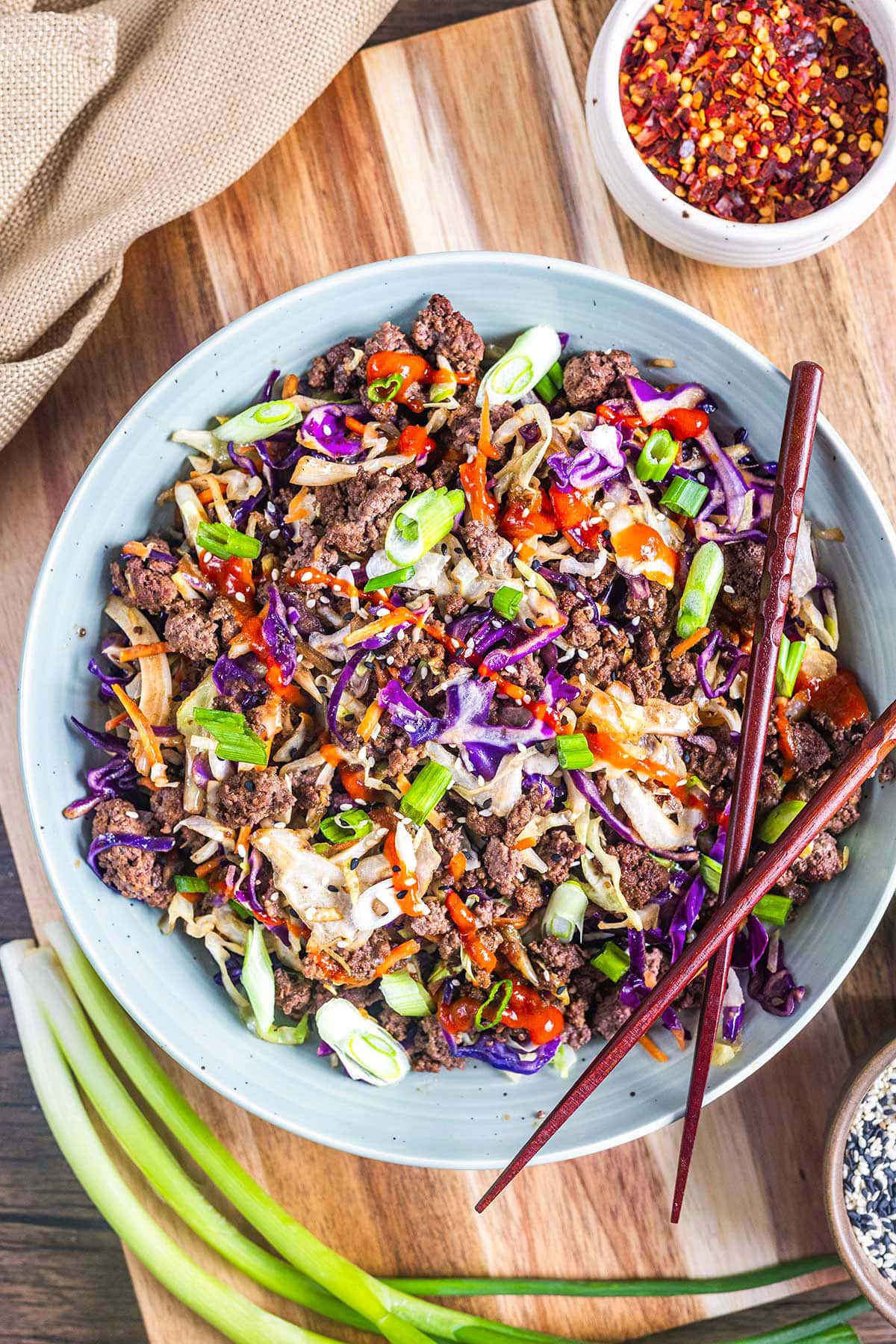 A vibrant keto egg roll in a bowl features ground beef, shredded cabbage, carrots, purple cabbage, and green onions with a drizzle of red sauce. Garnished with chopsticks, red pepper flakes and fresh green onions sit nearby on a wooden surface.