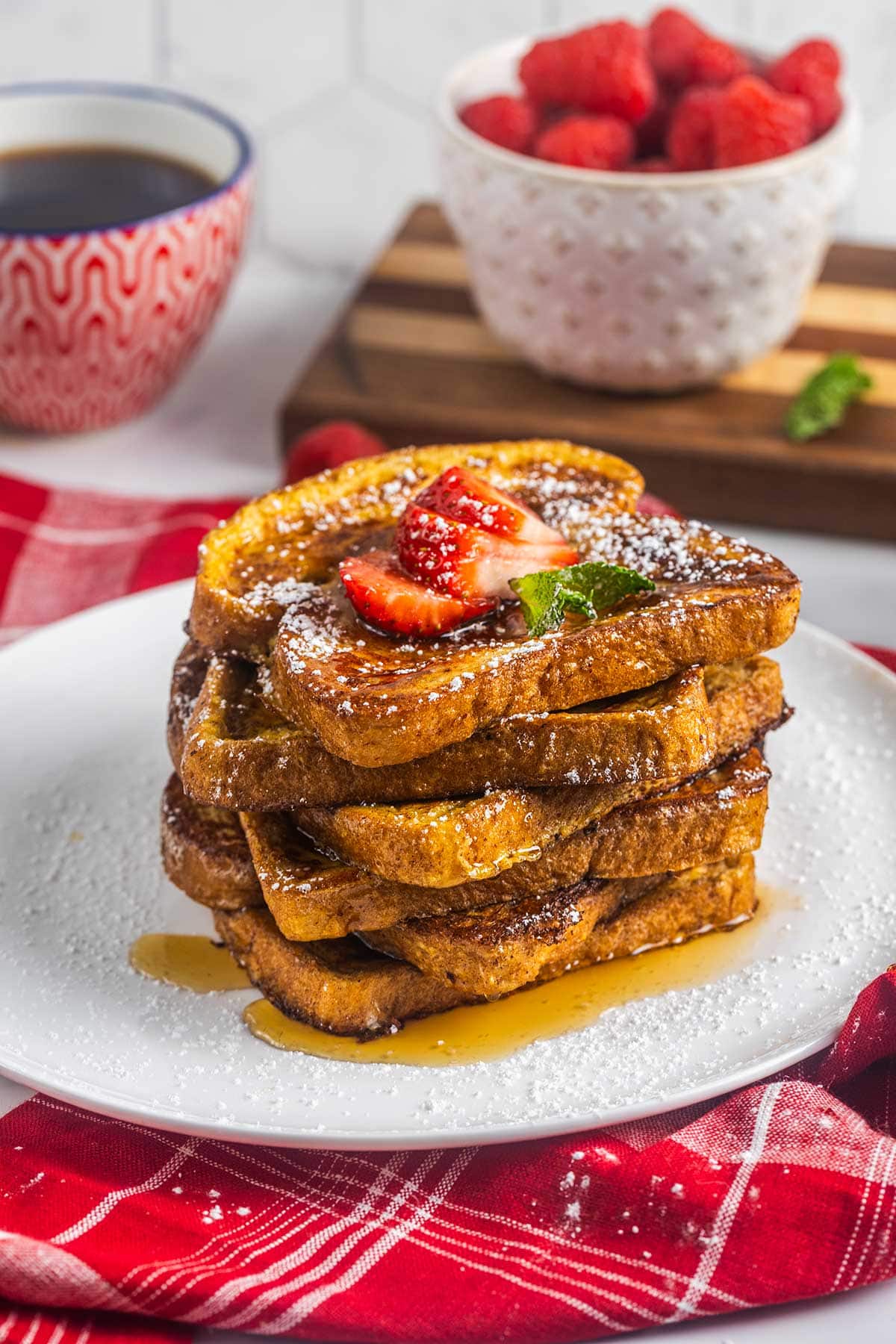 A stack of French toast topped with sliced strawberries, powdered sugar, and syrup sits on a white plate, while a red napkin, a cup of coffee, and even a keto big mac bowl add flair to the inviting breakfast scene.