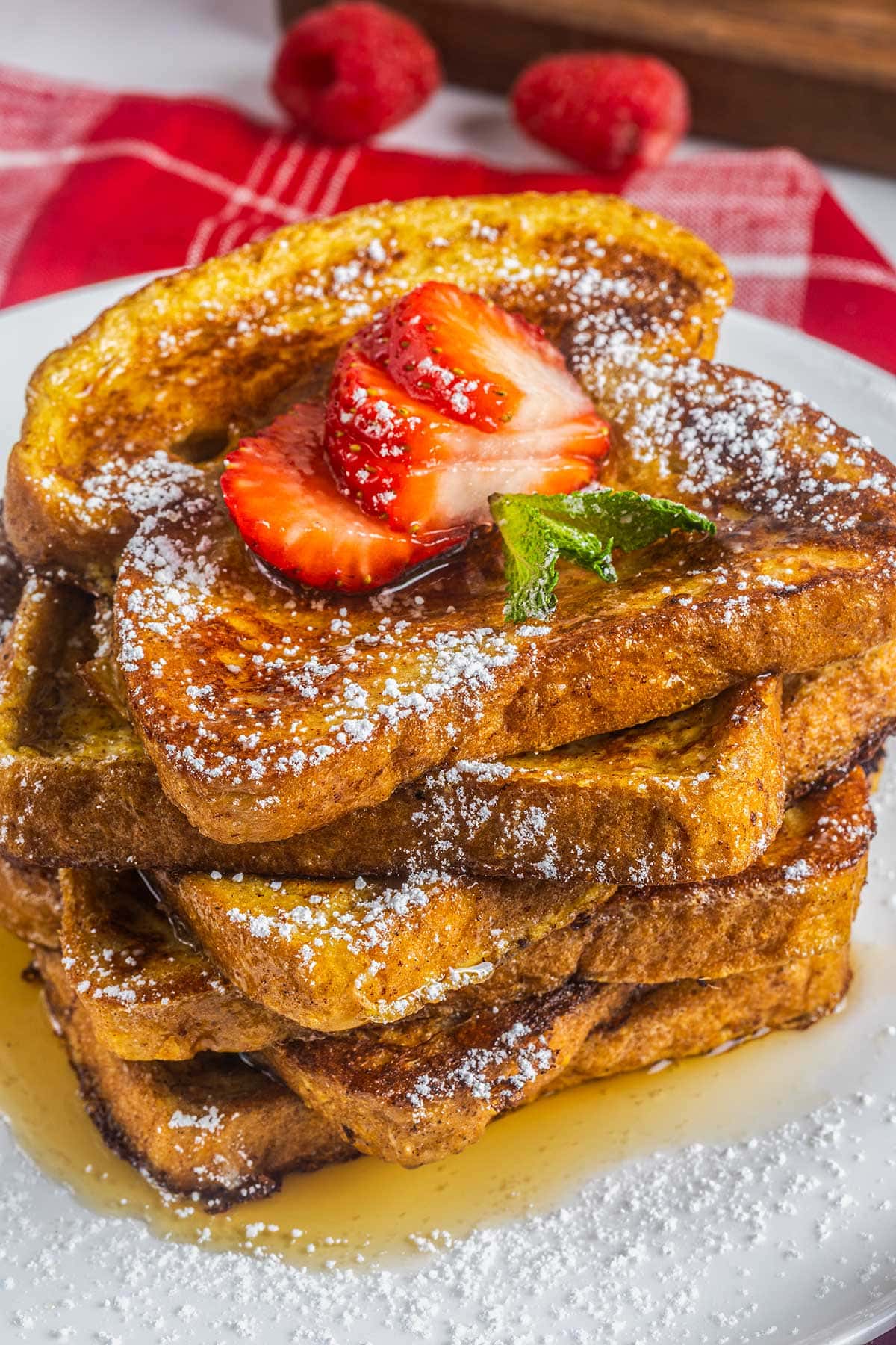 A stack of golden-brown French toast topped with sliced strawberries, mint leaves, powdered sugar, and syrup sits on a white plate—an indulgence that contrasts the savory flavors of a keto Big Mac bowl. Raspberries and a red napkin complete the scene.
