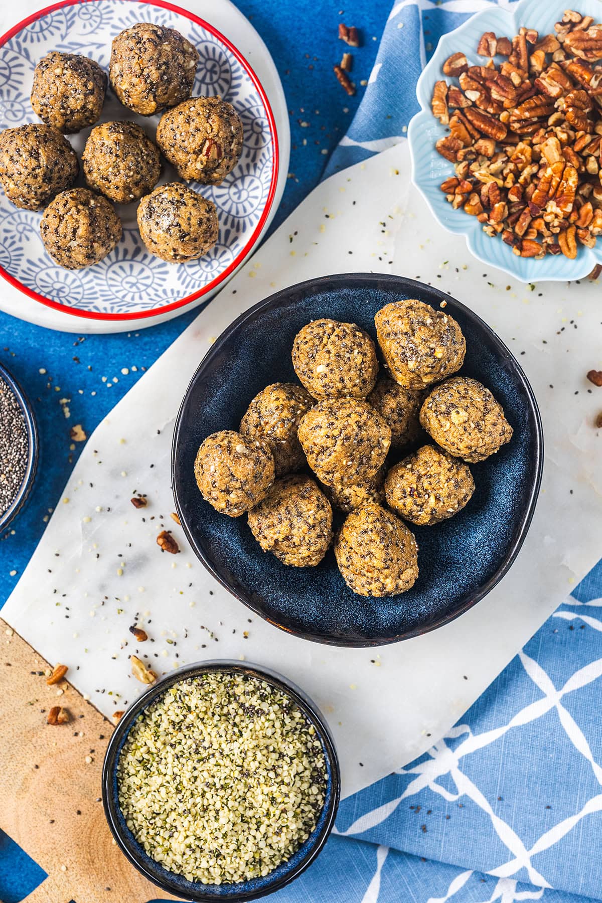 A blue plate with energy bites sits on a marble surface, surrounded by bowls of chia seeds, pecans, and hemp seeds, with more energy bites on a patterned plate nearby. A blue and white towel is partially visible.