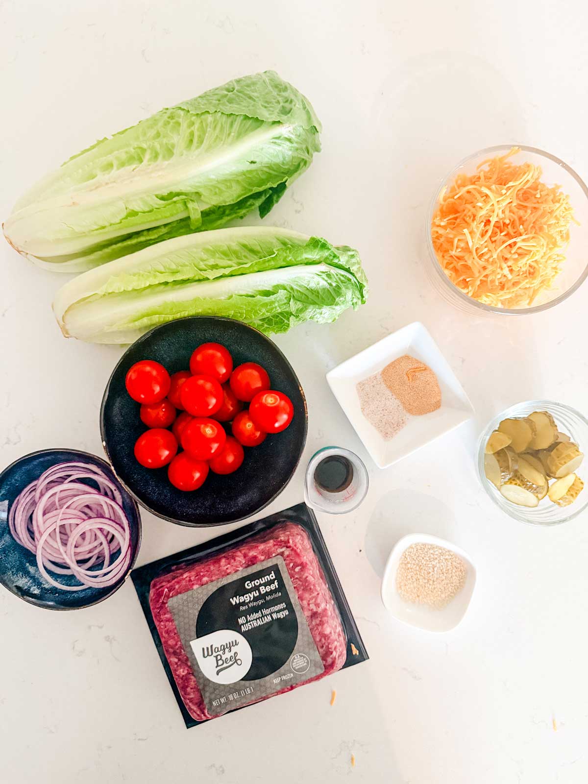 Ingredients on a white surface, including two romaine lettuce hearts, shredded cheese, grape tomatoes, sliced red onion, ground beef, pickles, seasonings, breadcrumbs, and Worcestershire sauce.