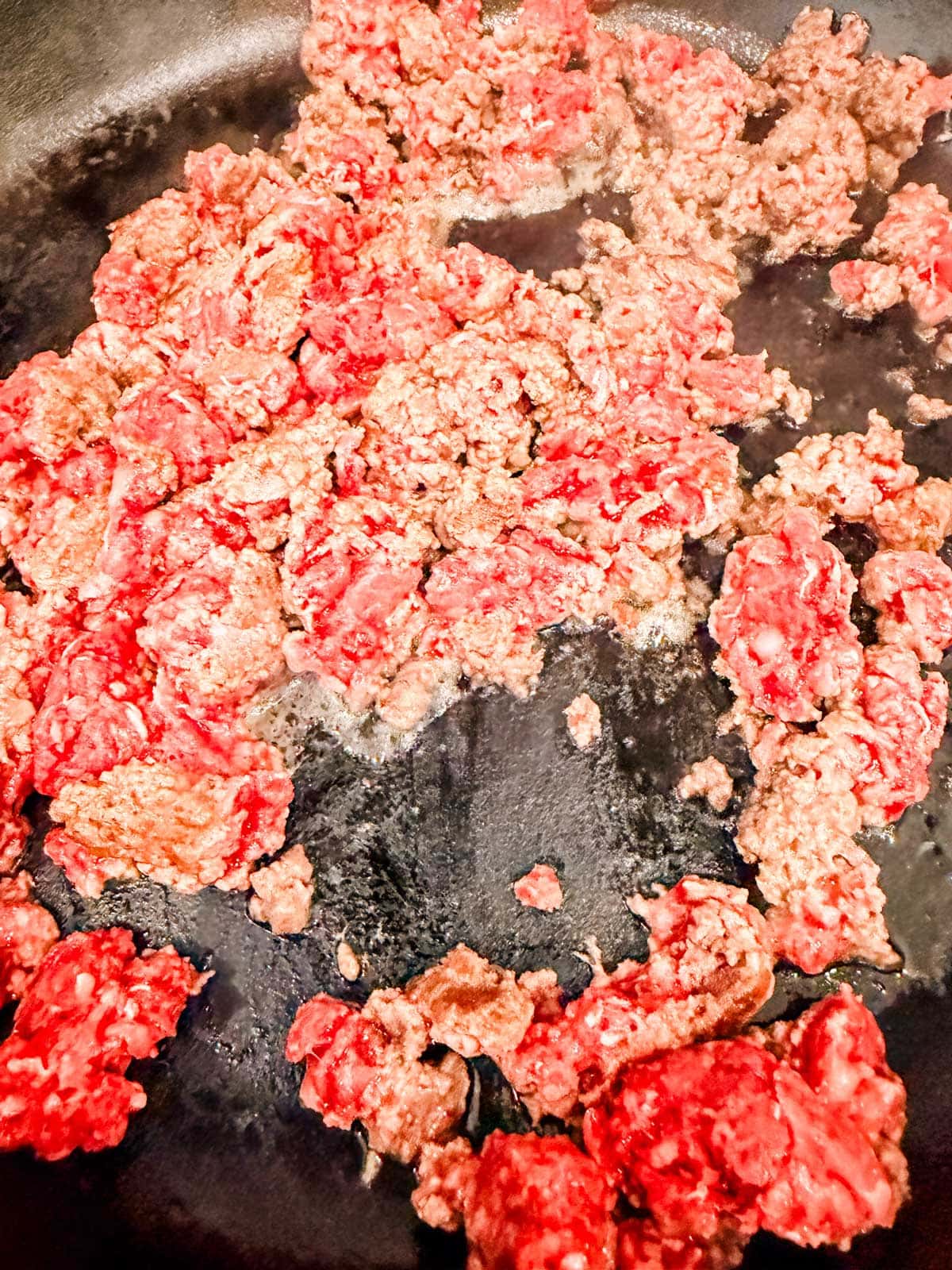 Ground beef being browned in a skillet, with some parts still red and raw while others are turning brown as they cook.
