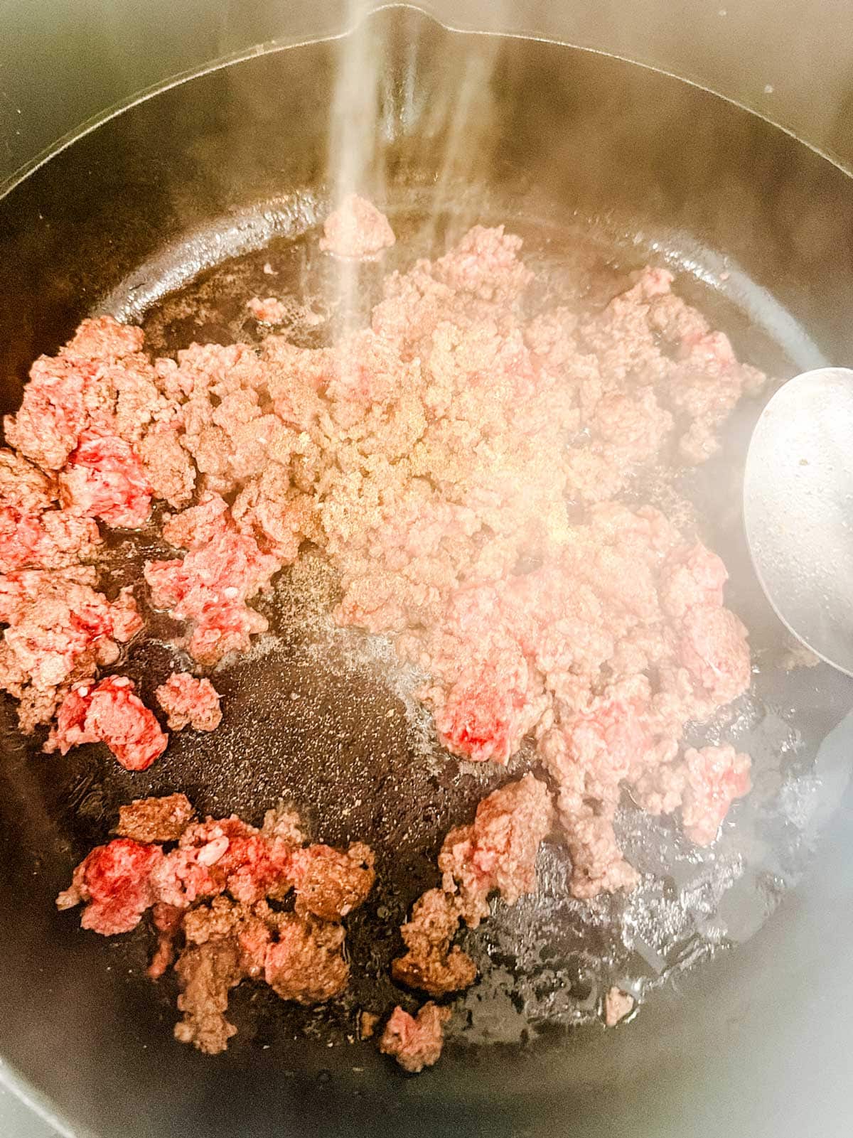 Ground beef is being browned in a cast iron skillet, with steam rising and a metal spoon resting on the side of the pan.
