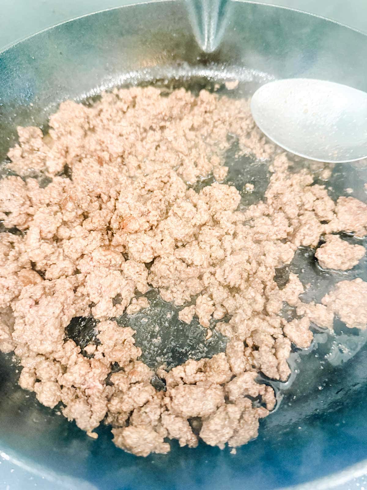 Ground beef cooking in a black skillet with a metal spoon visible on the right side, steam rising from the meat.