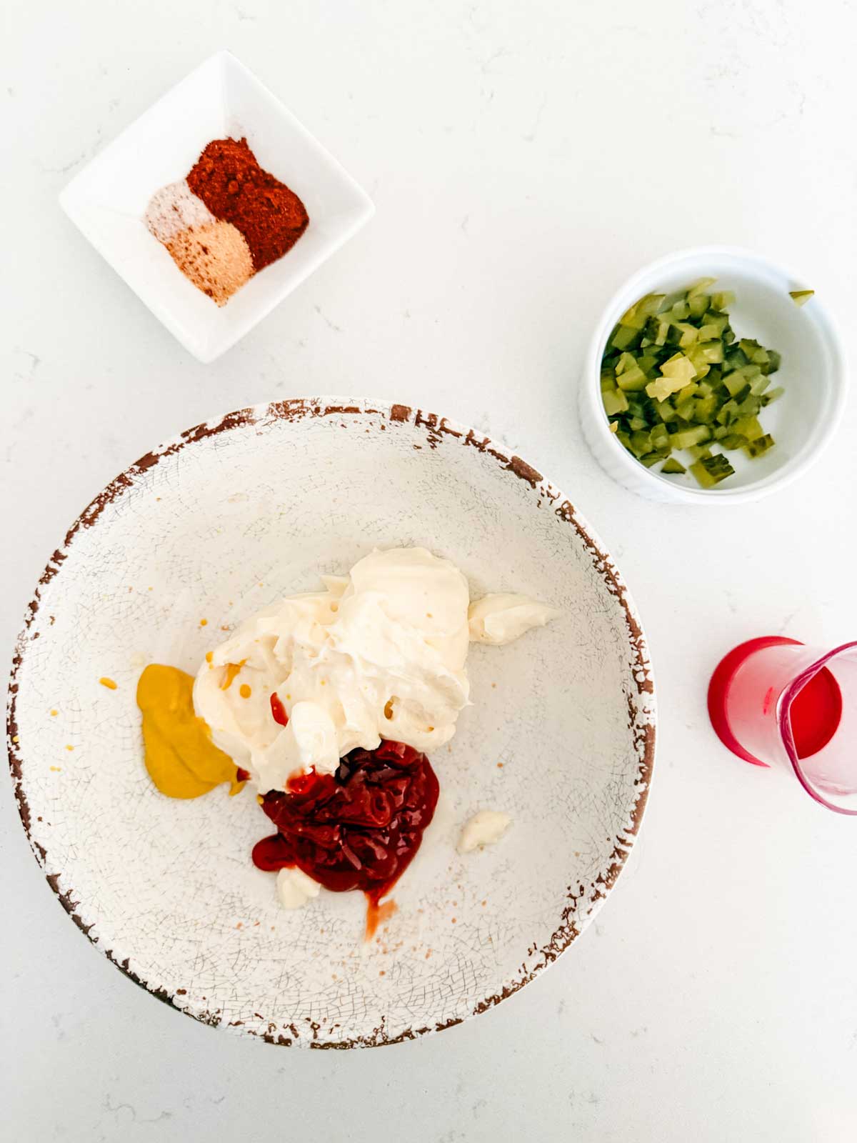 A bowl with mayonnaise, ketchup, and mustard, next to small bowls containing chopped pickles, seasonings, and a small amount of pickle juice on a white surface.