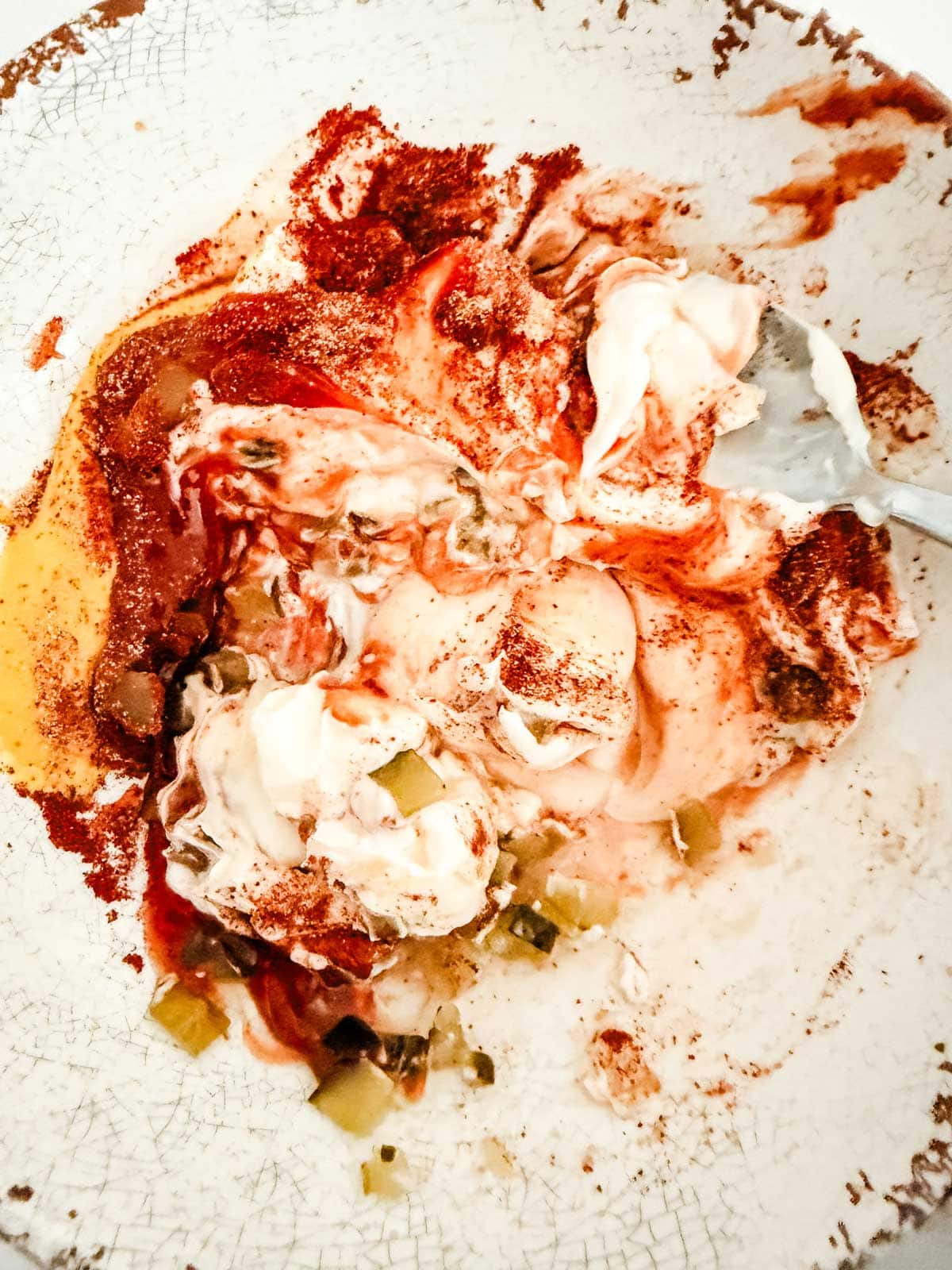 A bowl with a messy mixture of mayonnaise, ketchup, chopped pickles, and spices, partially stirred with a silver fork on the right side. The plate has a rustic, crackled texture and worn edges.