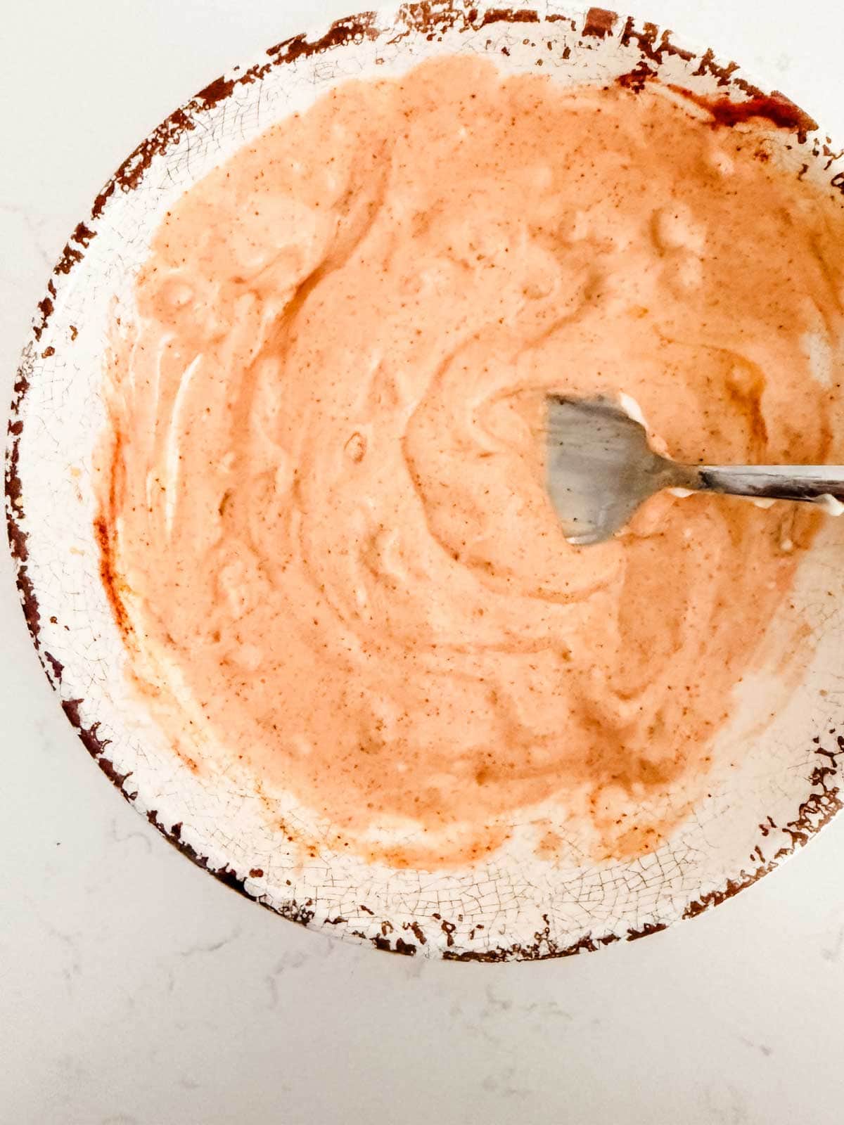 A ceramic bowl with a crackled glaze contains a creamy, light orange sauce or dip. A metal fork rests inside the bowl, which is set on a white surface.