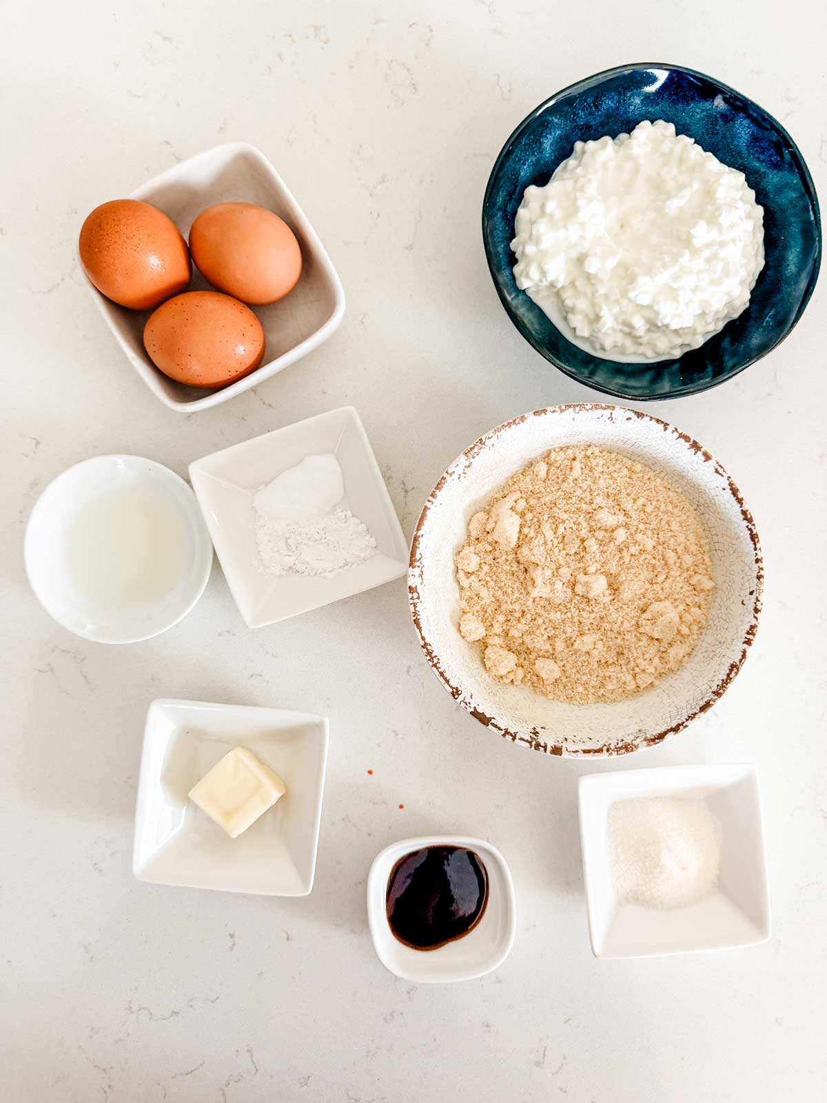 A top-down view of ingredients in separate bowls: three eggs, cottage cheese, almond flour, a pat of butter, a small bowl of sweetener, a small bowl of vanilla extract, baking powder), and a cup of liquid.