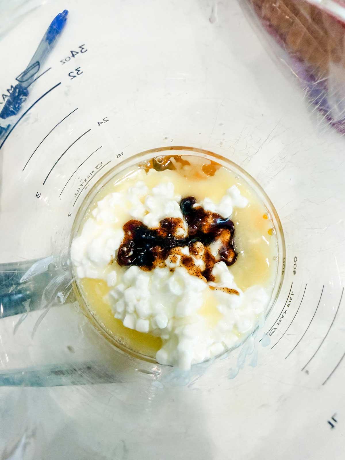 A top-down view of a measuring cup containing cottage cheese, a brown liquid seasoning or sauce, and a yellowish liquid. Measurement markings are visible on the cup, and a pen is partially seen in the background.