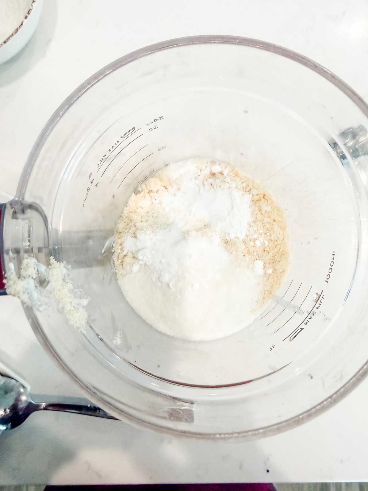 A clear mixing bowl containing white and beige dry ingredients sits on a white countertop, with a metal spoon resting nearby. Measurement marks are visible on the bowl.