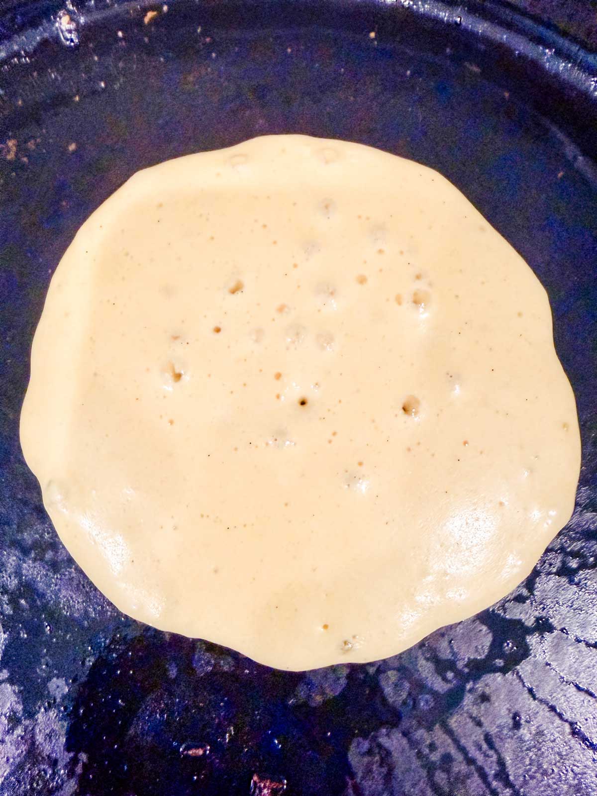 A pancake cooking on a dark skillet, with bubbles forming on its surface indicating it is being cooked on one side.