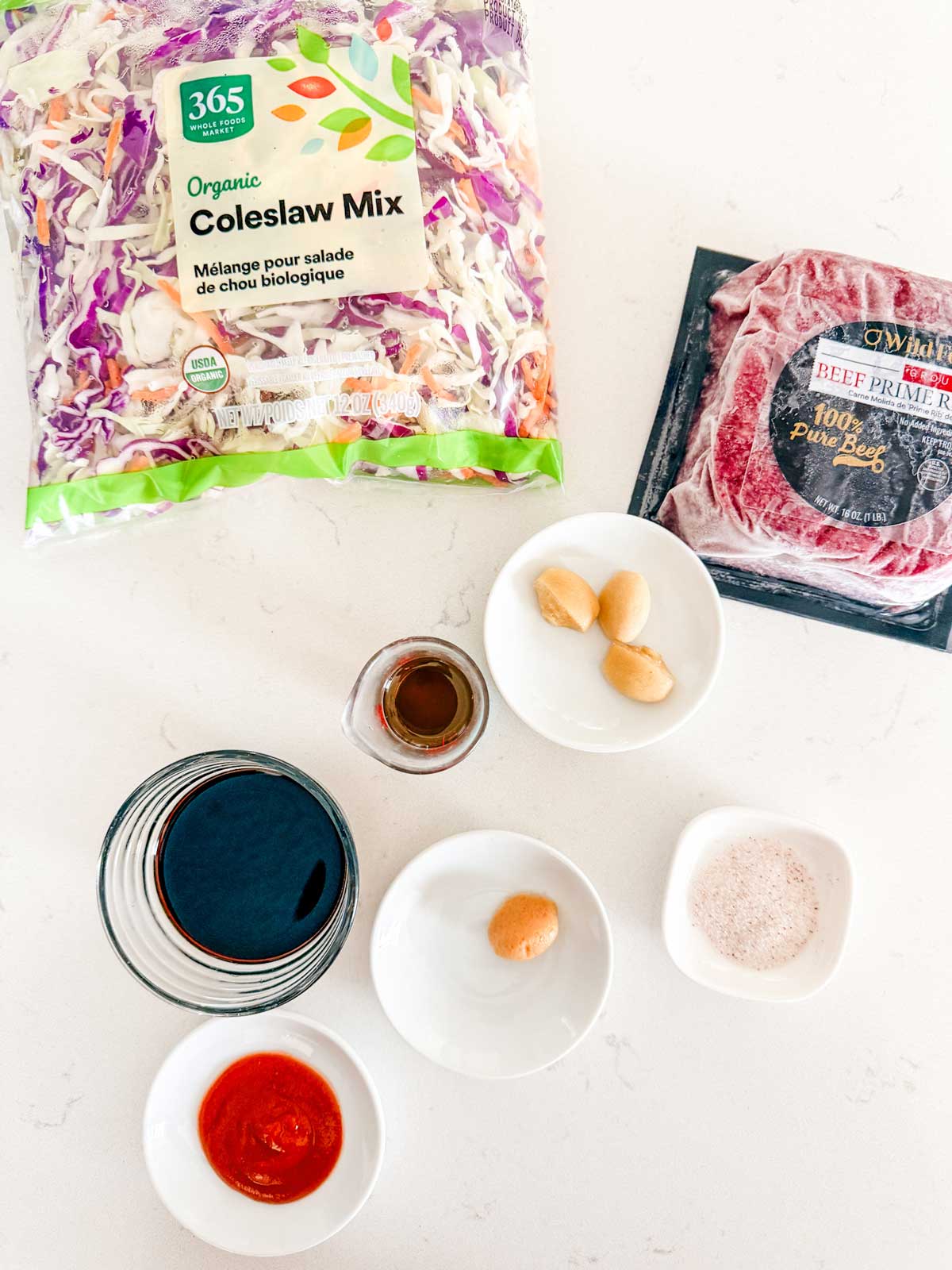 Ingredients on a white surface: a bag of organic coleslaw mix, a package of beef, three garlic cloves in a bowl, a bowl of coconut aminos, a bowl of sriracha, a bowl of salt, ginger and a small bowl of sesame oil.