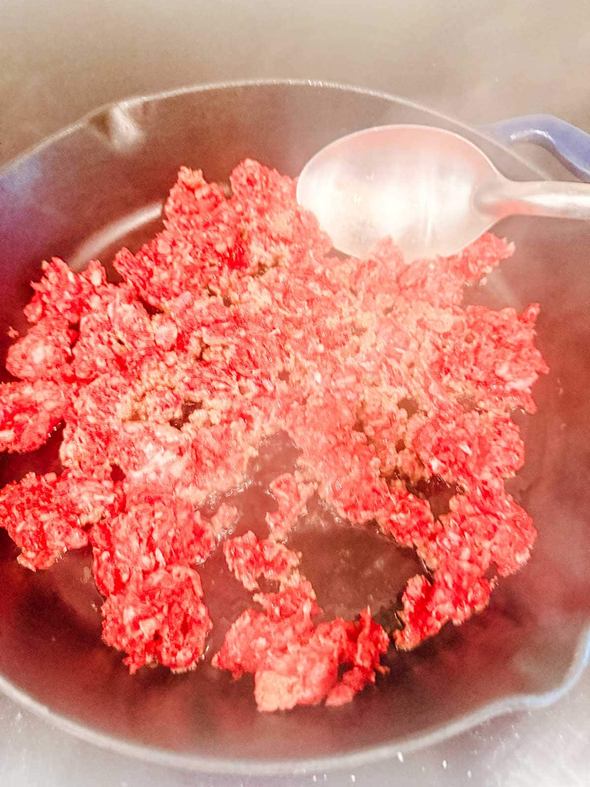 Ground beef browning in a black skillet with a metal spoon resting on the side, steam rising as the beef cooks.