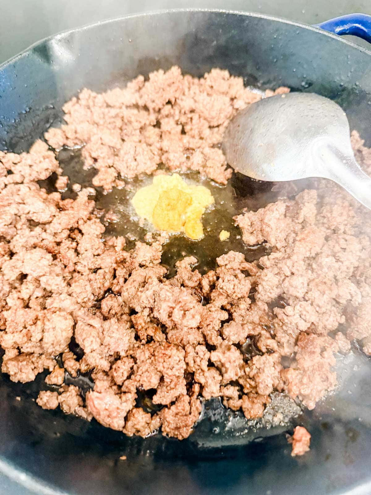 Ground beef cooking in a black skillet with a dollop of garlic and ginger in the center, and a metal spoon resting on the side. Steam is rising from the hot pan.