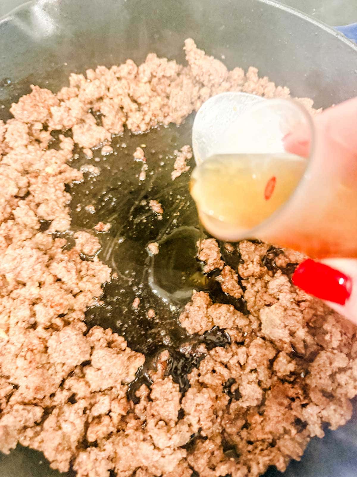 Ground beef cooking in a skillet as someone pours liquid from a small glass container into the pan. The person’s nails are painted red.